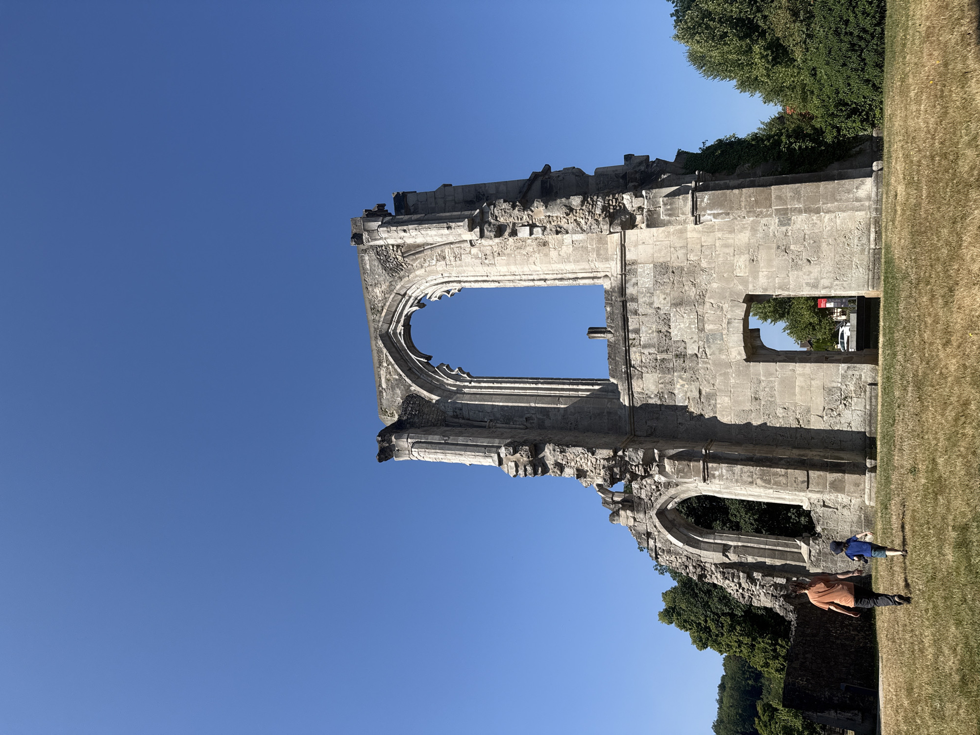 In Walkenried, Germany, two individuals, an adult and a child, are walking across a dry, grassy field towards the imposing stone ruins of a building, possibly a former abbey or church. The ruins are characterized by large, arched windows and crumbling stone walls, indicative of significant age and disrepair. The sky above is a clear, vibrant blue. Lush green trees and foliage flank the ruins, with a section of a stone wall visible on the left side of the frame. The scene is bathed in natural daylight under a cloudless sky.
