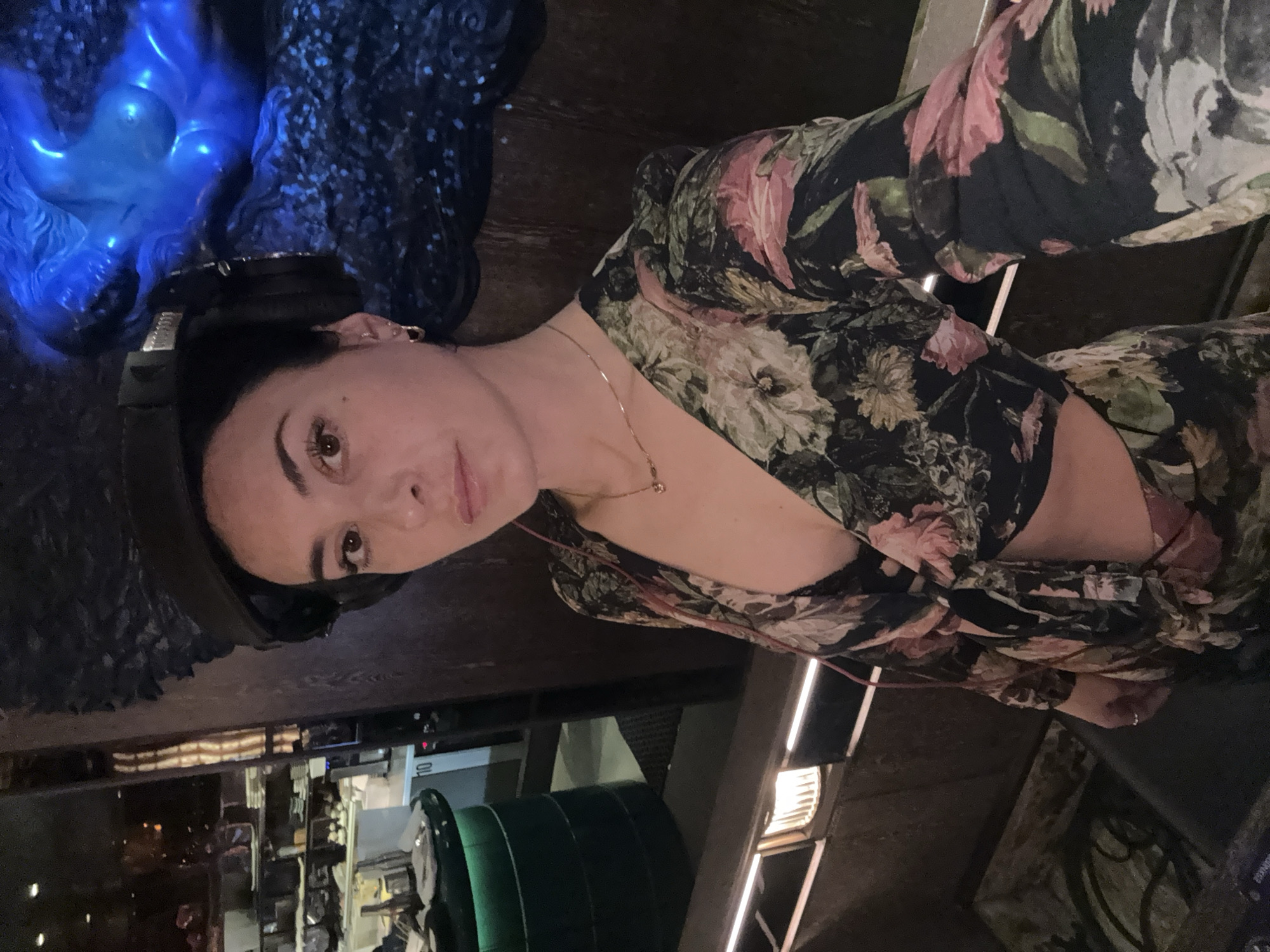 A woman with dark hair and light skin is posing for a selfie. She is wearing headphones and a floral-patterned dress with a deep V-neck. A thin gold necklace adorns her neck. Behind her is a blurred background, partially consisting of a blue illuminated sculpture and an out-of-focus interior of what appears to be a restaurant or bar. Elements of the kitchen or bar area are visible in the lower left corner, including stainless steel surfaces and green objects. Vertical light fixtures are also visible in the background. The location appears to be a restaurant interior based on the blurry background. Based on the unique blue sculpture, the location could be the restaurant "Park Row" in London. No specific interactions or actions are visible besides the selfie.