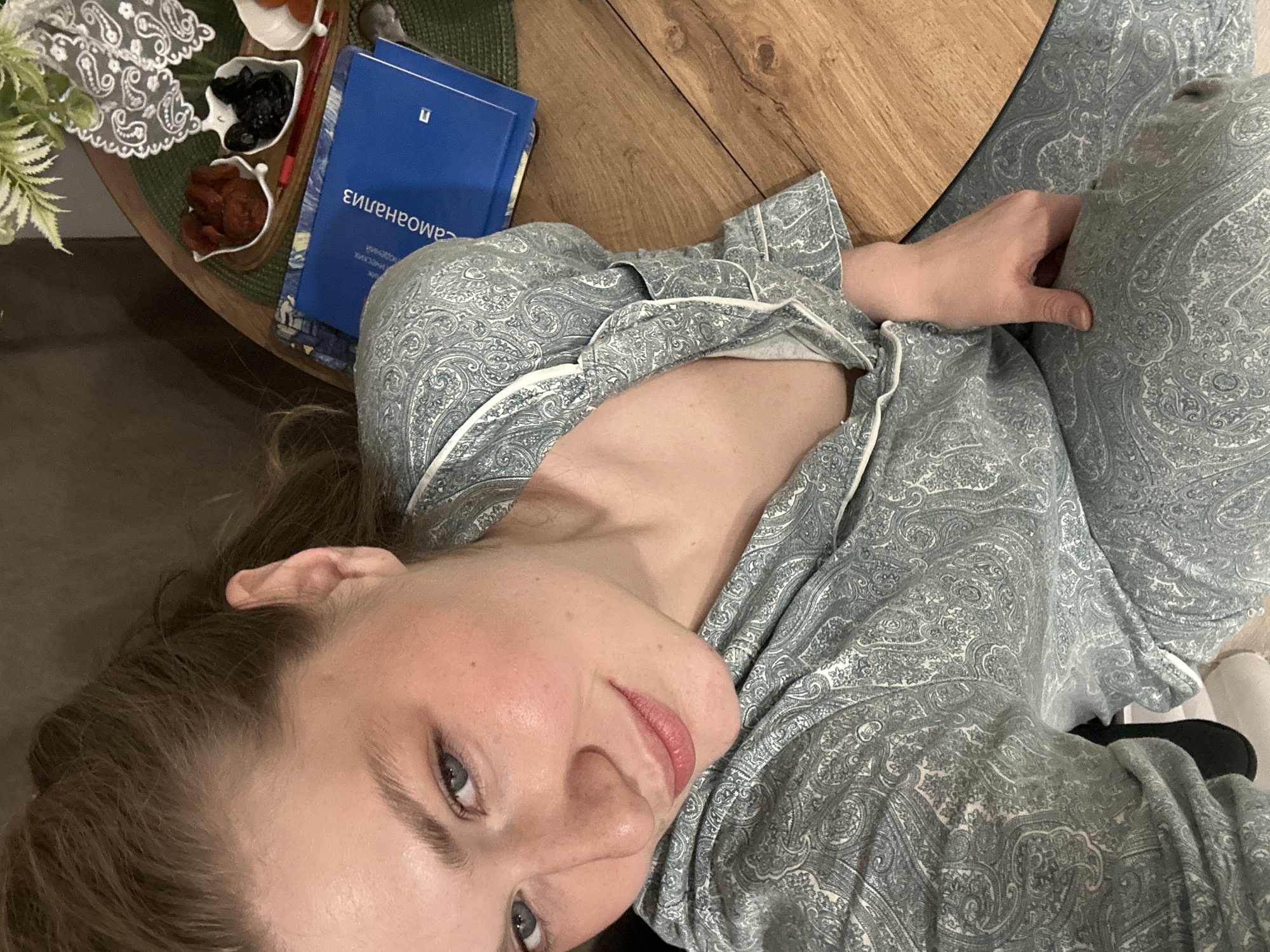 A selfie taken indoors shows a woman in a paisley-patterned pajama set. She is close to a round wooden table. On the table is a small tray with what appears to be dried fruit and a blue book with text in Russian. The lighting is soft, suggesting natural daylight. The location is likely inside a building, but there is no verifiable evidence to suggest this scene is in Kazan, Russia.