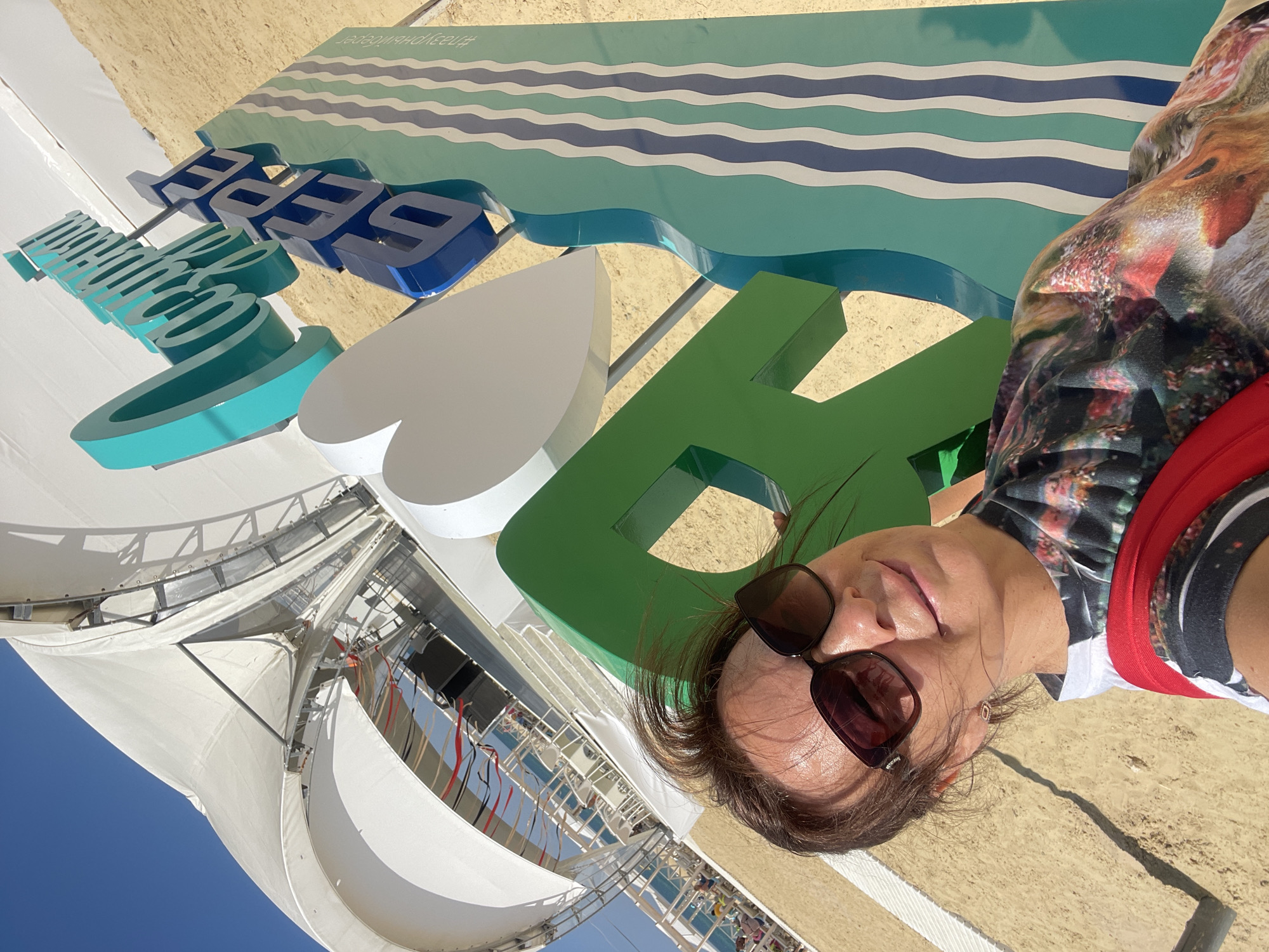 A woman wearing sunglasses and a t-shirt with a colorful print takes a selfie in front of a large, blue and green sign that says "ЛЕТО" (Summer) and "БЕРЕГ" (Shore). The sign is decorated with a heart and wavy lines. Behind the sign, there is a large white tent structure, likely part of a beach resort or entertainment venue. The ground is covered in sand, indicating a beach environment. In the background, the sea and a clear blue sky are visible. The location is identified as the city of Anapa, Russia.