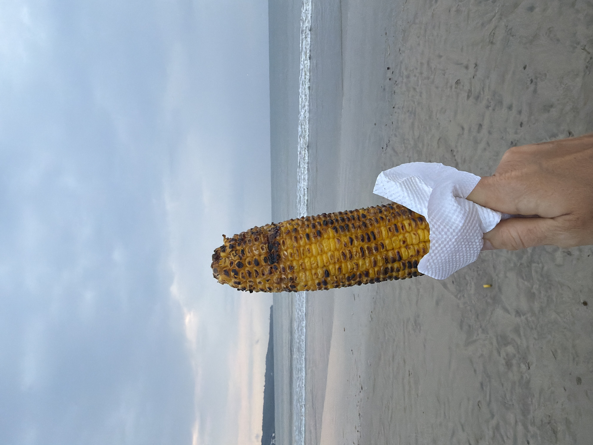 A human hand, light-skinned, is positioned in the lower right, holding a grilled corn on the cob. The corn is primarily yellow with distinct dark brown and black charred kernels. It is wrapped at its base by a white, textured paper napkin. The foreground consists of a light gray-brown sandy beach with subtle indentations. Beyond the sand, the ocean extends into the midground, featuring gentle, white-capped waves breaking near the shore. A dark, low-lying landmass is visible on the far left horizon. The sky is overcast with light gray cloud formations. The scene is Jimbaran Beach in Jimbaran, Indonesia.