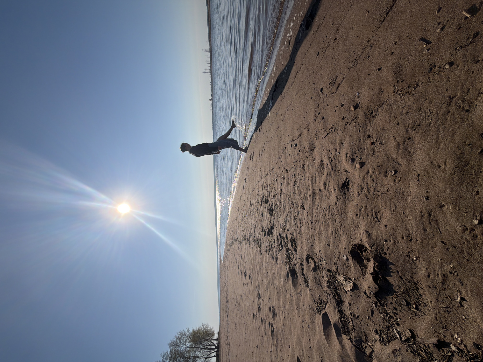 A man in dark clothing is standing at the edge of a body of water, possibly a sea or large lake. The sun is bright in the sky, causing glare. The man's left leg is raised. The scene is on a sandy beach, with the water meeting the sand. In the background is a structure, likely an industrial or port facility. A small tree is visible in the bottom-left corner of the image. The scene is in Noviy, Russia.