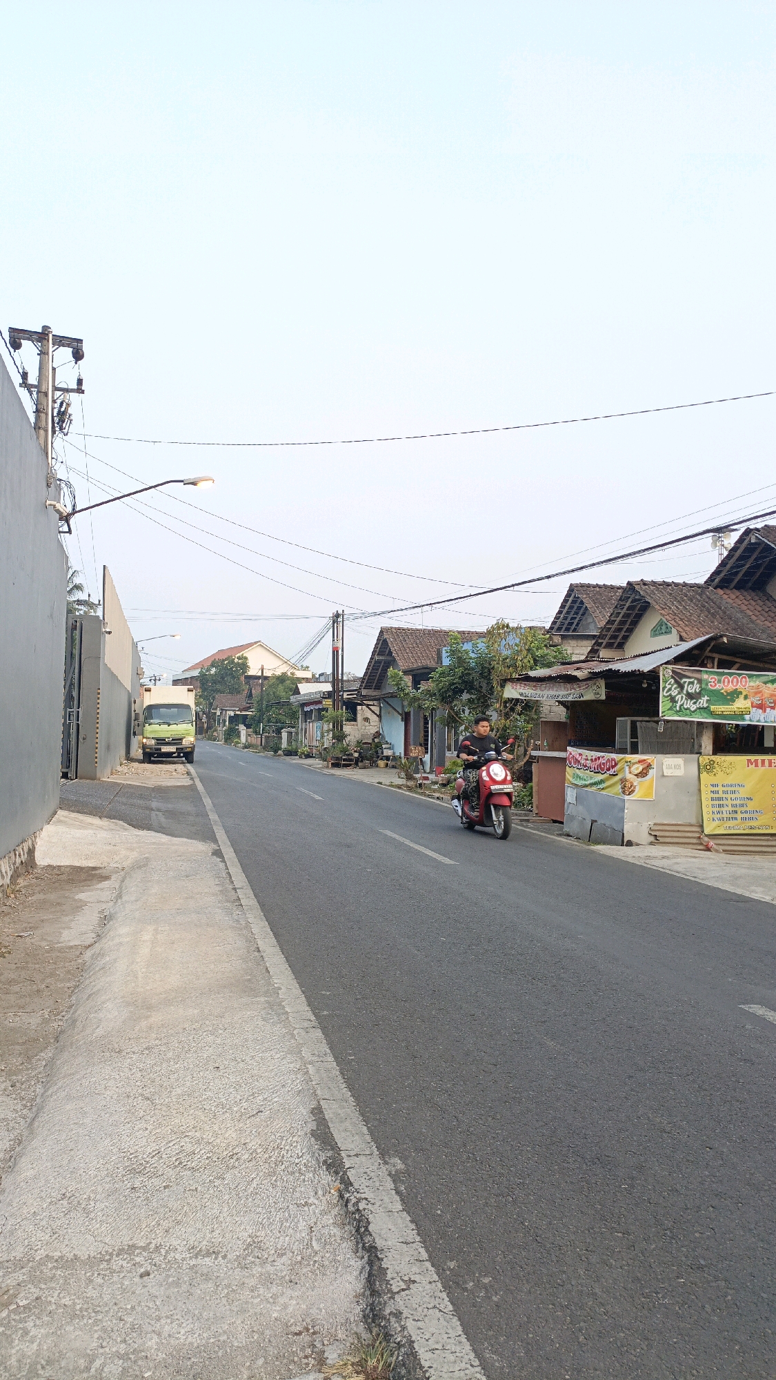 This is a full-frame, eye-level, vertical, outdoor photograph taken during the day on a paved road in Salatiga, Indonesia. The road occupies the majority of the image, with a concrete sidewalk on the left and buildings lining the right. A person is riding a red scooter towards the viewer on the right side of the road. The rider is wearing a dark t-shirt and appears to be male. Behind the scooter, there are several low-rise buildings with pitched roofs, some appearing to be businesses with signs. One sign, in the upper right, displays "Es Teh Pusat 3.000" and what appears to be an advertisement for food. Further down the road on the left, a light green truck is parked. Overhead, power lines and streetlights are visible against a pale sky. The overall scene depicts a typical street in a moderately urban or suburban area of Salatiga, Indonesia.