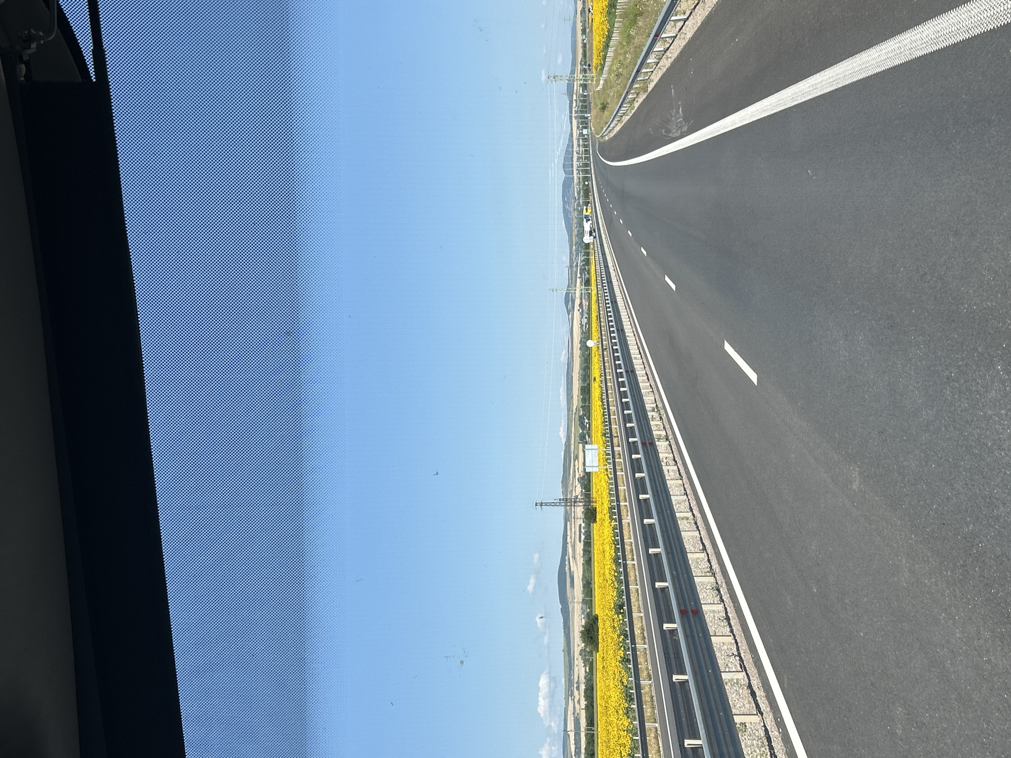 The image depicts a highway with a sunflower field to the left and right, stretching towards a hazy horizon. The highway has two lanes in the visible direction, separated by a dashed white line. A guardrail runs along the edge of the highway. The sky is clear and blue. The scene appears to be a rural or semi-rural area. There is no visible indication of the city of Prolesha, Bulgaria, in the image.