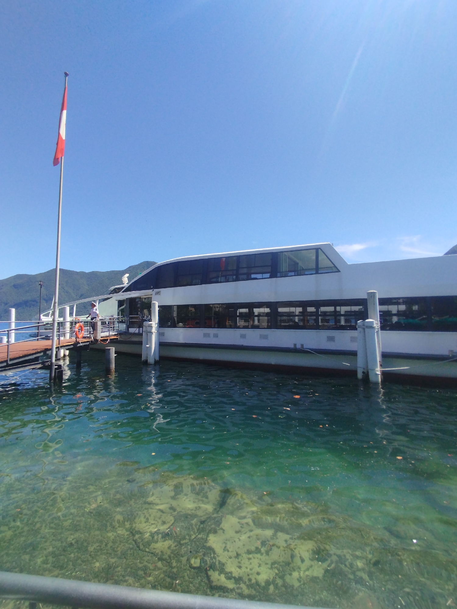 The image depicts a modern white passenger boat docked at a pier on a clear day. The boat has large windows along its side and a distinctive angled window at the front. A person in a white uniform and cap stands on the wooden pier, near an orange life preserver. A Swiss flag on a tall flagpole is visible to the left of the boat. The water is a vibrant turquoise, with visible details of the lakebed below. In the background, lush green hills rise under a bright blue sky. The scene appears to be at a lakeside promenade or port in Lugano, Switzerland, given the flag and the surrounding landscape.