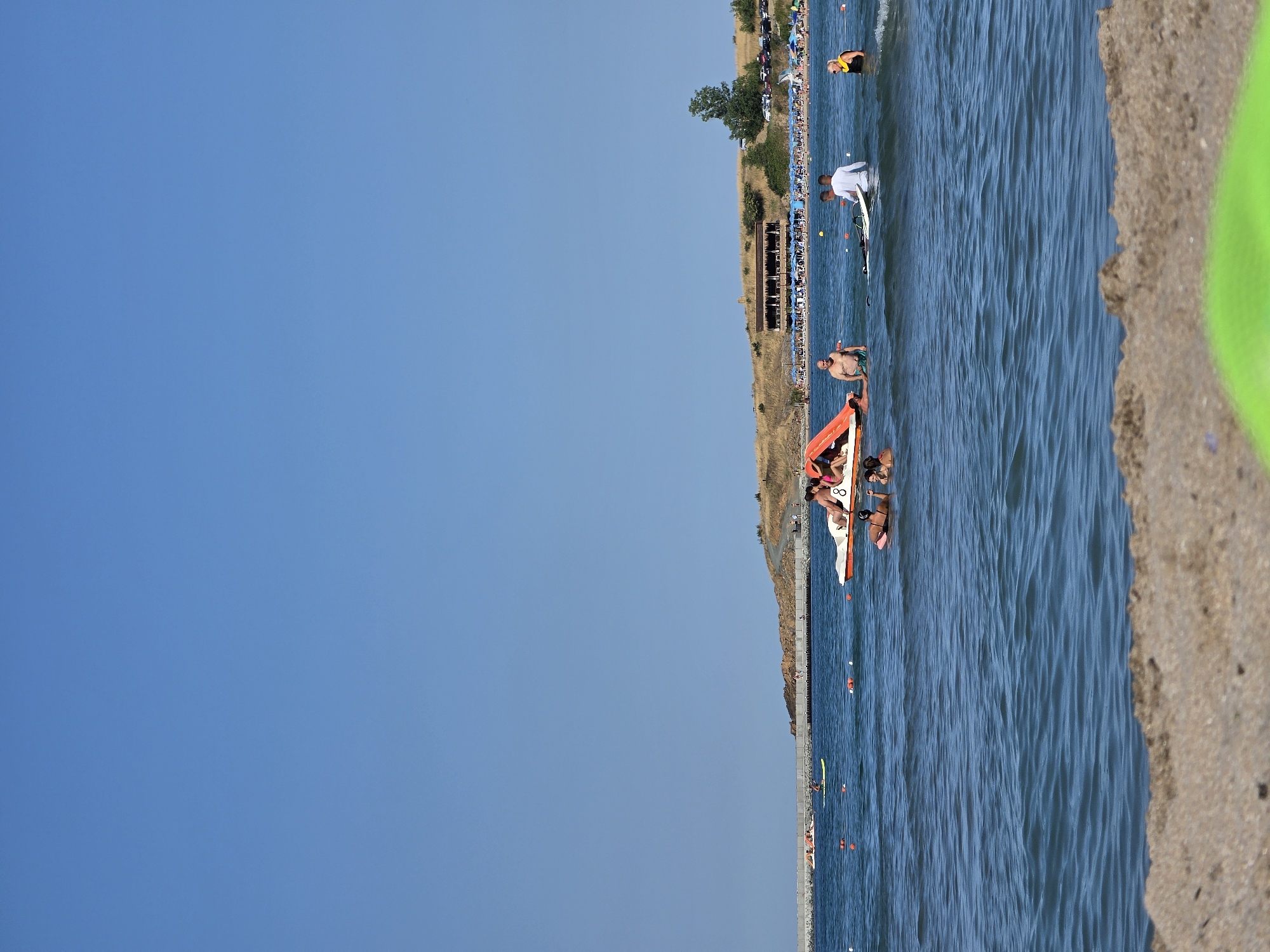 This is a vertical, outdoor, full-shot photograph taken on a sunny day at a beach in Ahtopol, Bulgaria. The foreground shows a sandy beach with a bright green object partially visible at the bottom. The midground is a blue, slightly wavy sea populated by several people. In the center-left of the water, a group of people are on a bright orange inflatable slide attached to a boat. To the right of this boat, two men are standing in the water, one holding a surfboard. Further to the right, a woman wearing a yellow life vest is in the water. In the background, a long stone pier stretches across the sea. Beyond the pier, there is a hillside covered in dry grass and vegetation. On the hillside, a building with a thatched roof is visible, and behind it, rows of blue umbrellas indicate a beach with sunbathers. A line of parked cars is also visible on the hillside. The sky above is clear and blue.