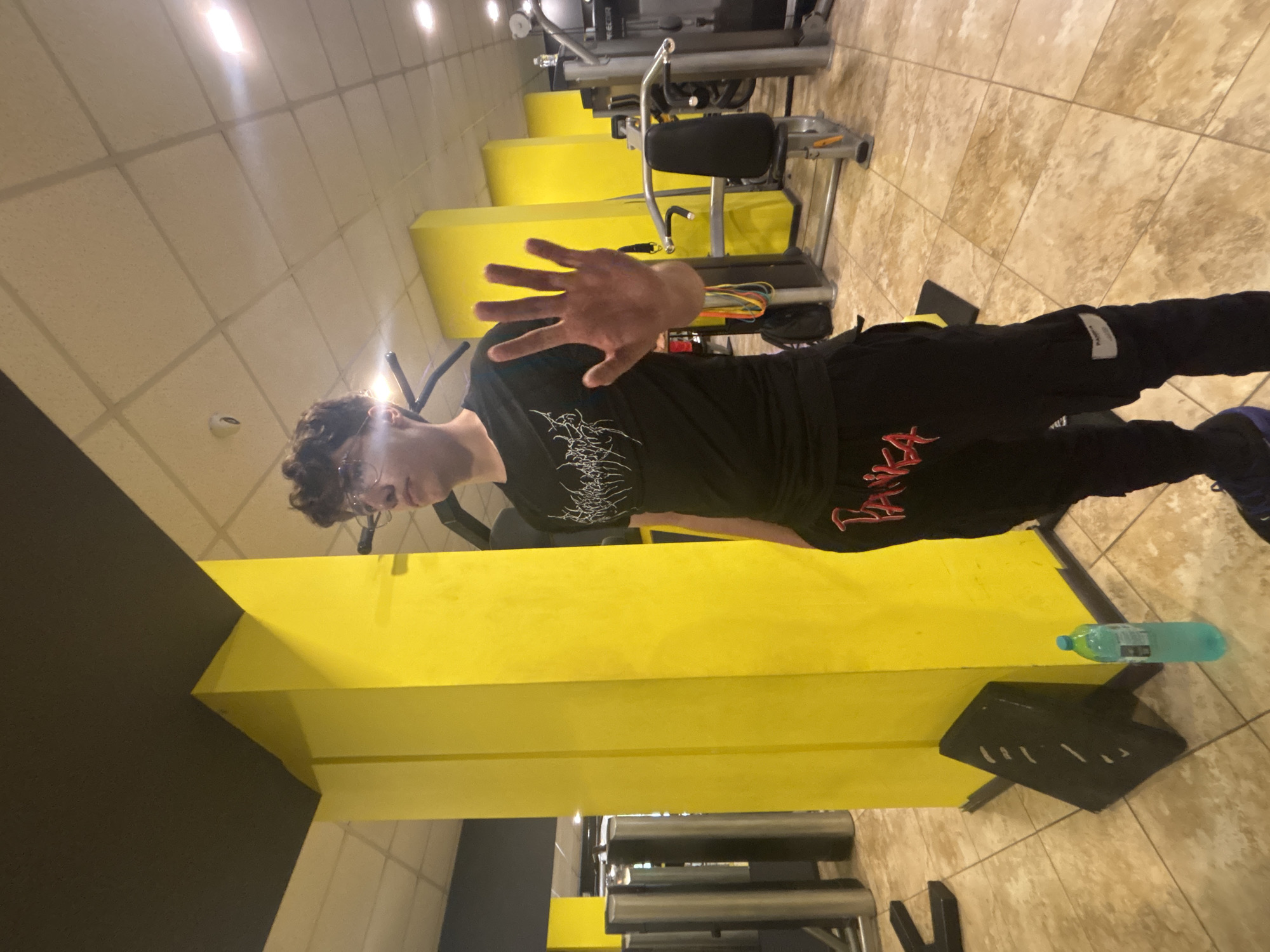 A young, light-skinned person with curly brown hair and glasses is standing in a brightly lit gym with tan tiled flooring. They are wearing a black t-shirt with white graphic lettering and black pants with red graphic lettering. Their right hand is raised, palm facing forward, towards the camera. The gym features yellow support pillars and exercise machines. A blue water bottle rests on the floor near a black weight support. The location is presumably a gym in the city of Troyan, Bulgaria.