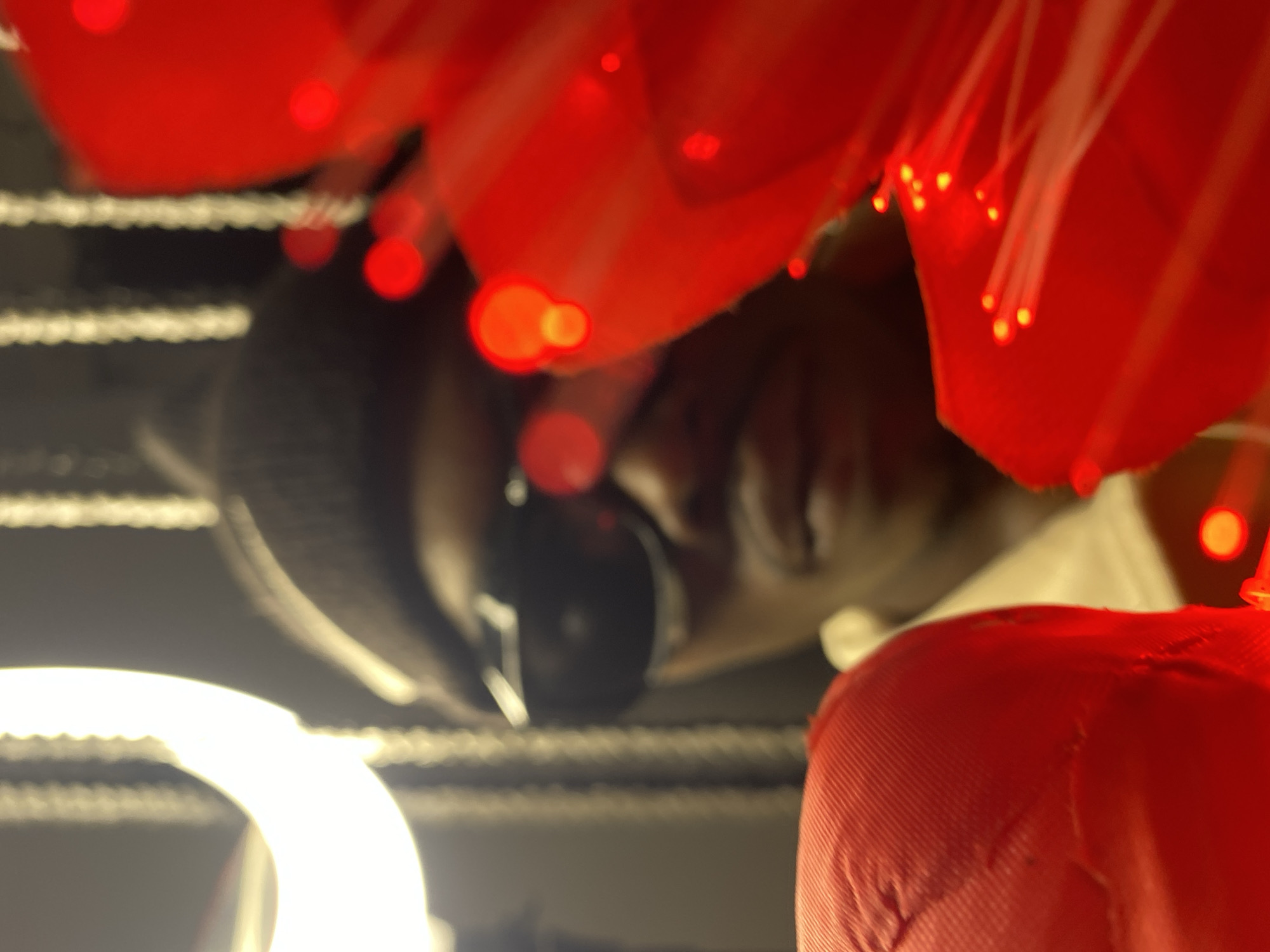 The image is a close-up, low-angle, blurred, self-portrait of a dark-skinned person wearing a black beanie, sunglasses, and a white shirt. The person is positioned in the center of the frame, with their face partially obscured by red elements in the foreground. To the left, a large, out-of-focus red object, possibly a hat, occupies the lower-left corner. To the right, a large, out-of-focus red object with small, bright lights, resembling a decorative flower with fiber optic lights, dominates the right side of the image. In the background, a bright, out-of-focus ring light is visible on the left, and vertical, blurry lights are present in the middle ground. The overall impression is one of artistic self-expression, possibly taken in an indoor setting with artificial lighting. The image does not contain any specific visual cues that would indicate its location in Abakaliki, Nigeria.