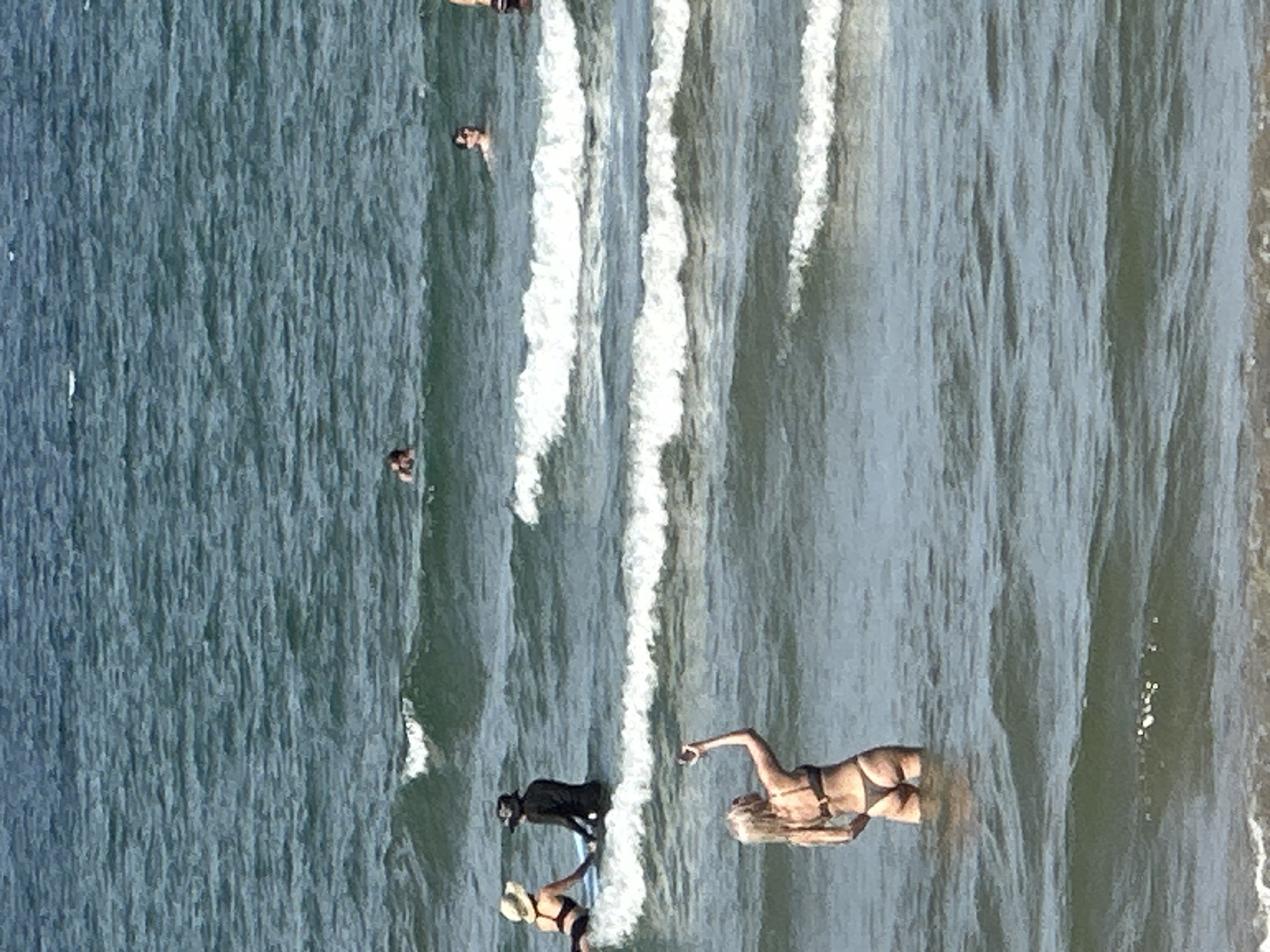 The image depicts a beach scene at Da Nang, Vietnam. In the foreground, a blonde woman in a bikini stands in the shallow water, holding a mobile phone up to take a selfie. In the mid-ground, two people are seated on a surfboard in the water. One person is wearing a hat and dark clothing, while the other wears a straw hat and a bikini. Several other individuals are visible in the water further out, with some swimming and others wading. The ocean is wavy, with whitecaps indicating moderate surf conditions. The sky is a pale blue, suggesting a sunny day.