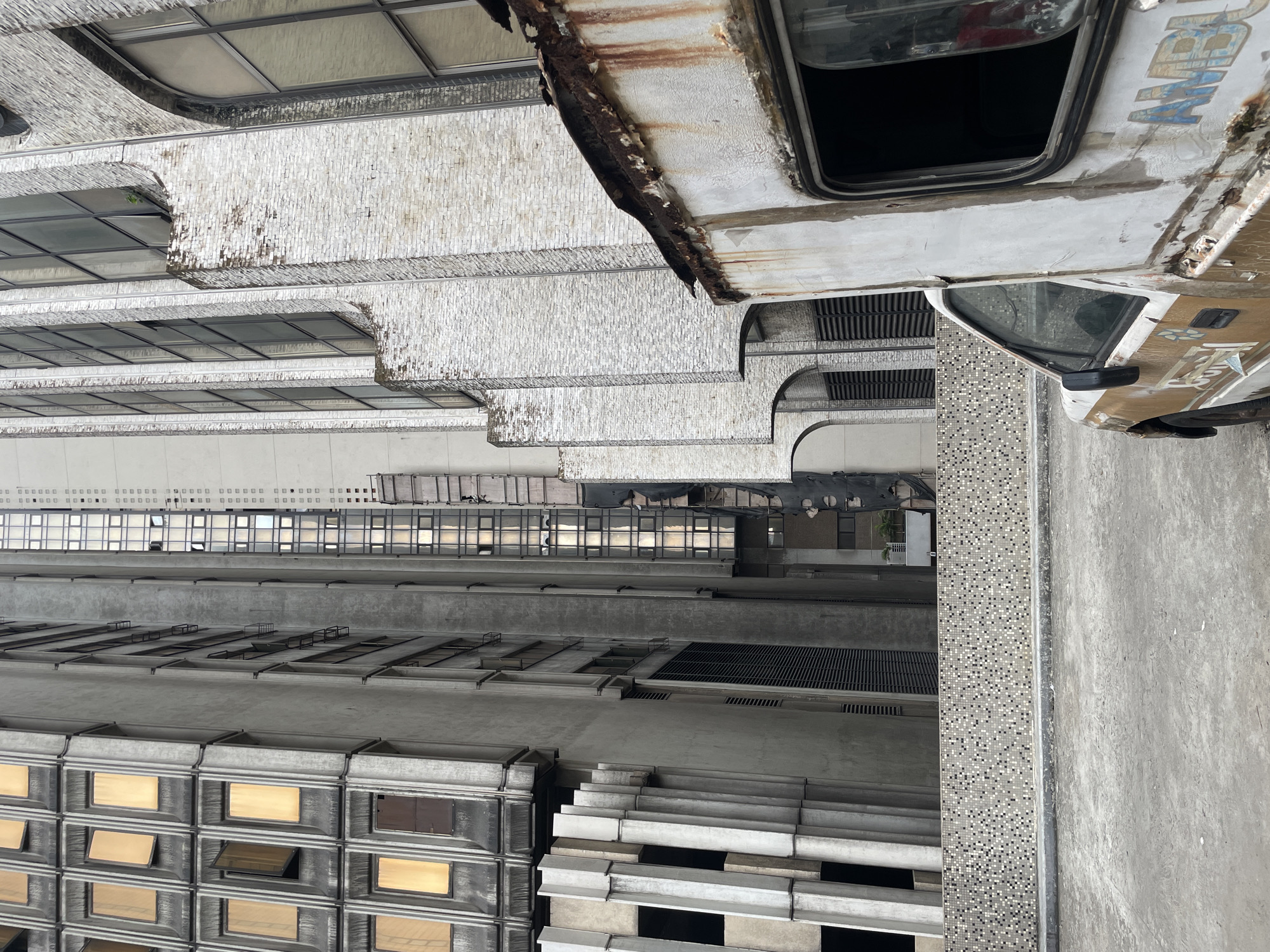 The image depicts a weathered, high-angle view of Lagos, Nigeria, likely taken from an elevated position. A derelict vehicle, possibly a van or small bus, is positioned in the upper right corner, exhibiting rust and deterioration. Adjacent to the vehicle and extending downward is a building with repetitive window patterns and tiered architectural elements, suggesting an office or residential structure. The architecture appears aged, with visible signs of wear and tear. Lower down, a concrete surface or walkway runs parallel to the building. The overall environment suggests urban decay or neglect.
