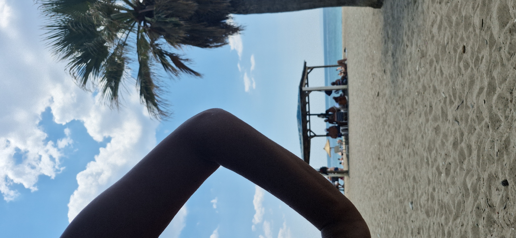 The image depicts a beach scene at the city of Paleo Faliro, Greece, under a partly cloudy sky. In the foreground, a silhouetted arm is visible, angled from the left side of the frame across the top portion. The middle ground features a sandy beach with a thatched-roof structure providing shade for several people sitting on chairs. Beyond this structure, the blue sea stretches to the horizon, with a few scattered beach umbrellas and more people visible along the shoreline. A palm tree with dense fronds is partially visible on the right side of the frame. The overall ambiance is that of a sunny day at the beach.
