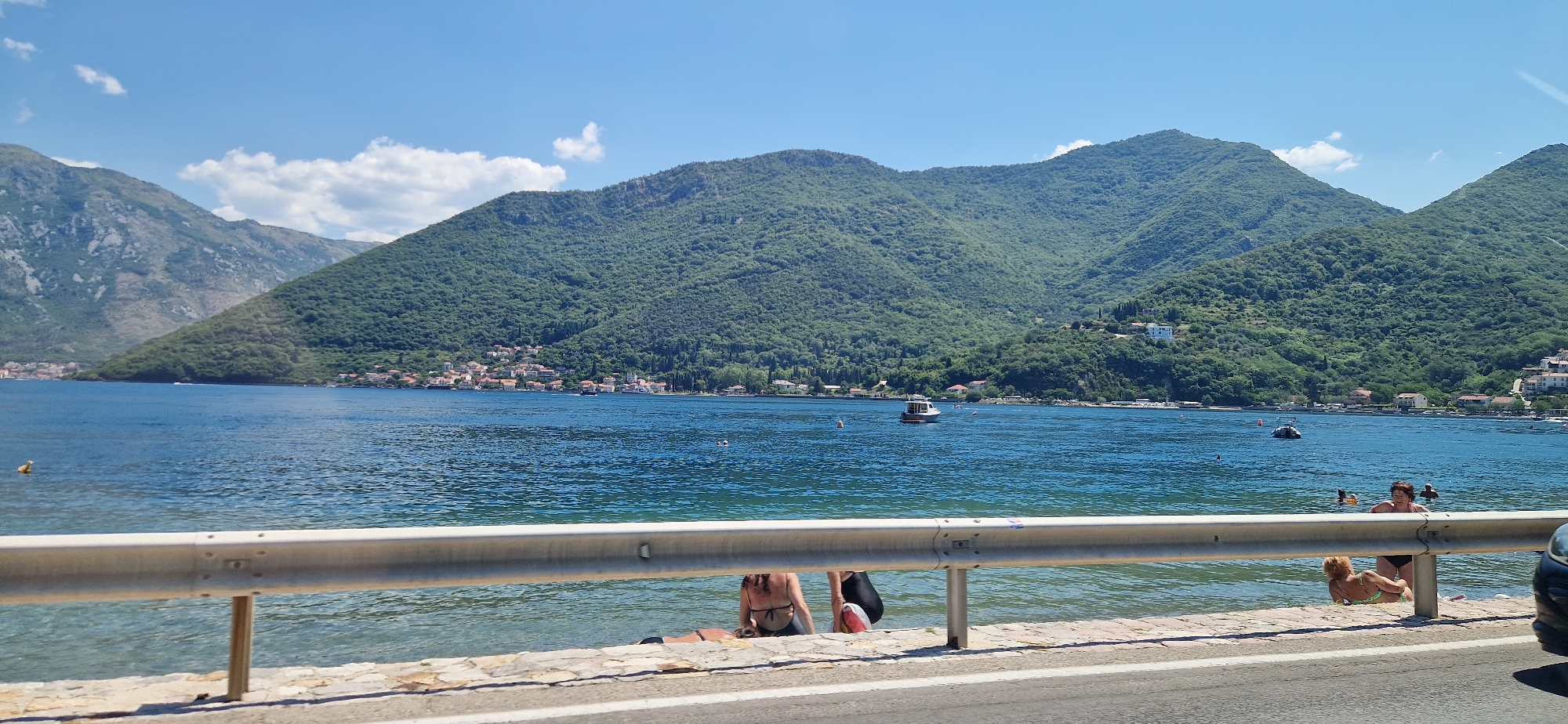 The image depicts a scenic view of the Bay of Kotor near Jošice, Montenegro. In the foreground, a metallic guardrail runs horizontally across the frame, with a stone embankment below it. Beyond the guardrail, the calm, blue water of the bay stretches towards a verdant, mountainous coastline dotted with buildings. Several people are visible in the shallow water near the shore, some appearing to be swimming or relaxing. A small boat is situated in the bay, and buoys are also present. The sky is clear with a few scattered clouds, and the sunlight illuminates the landscape. The mountains are covered in dense green foliage, suggesting a warm, temperate climate. The overall impression is one of a peaceful coastal environment on a sunny day.