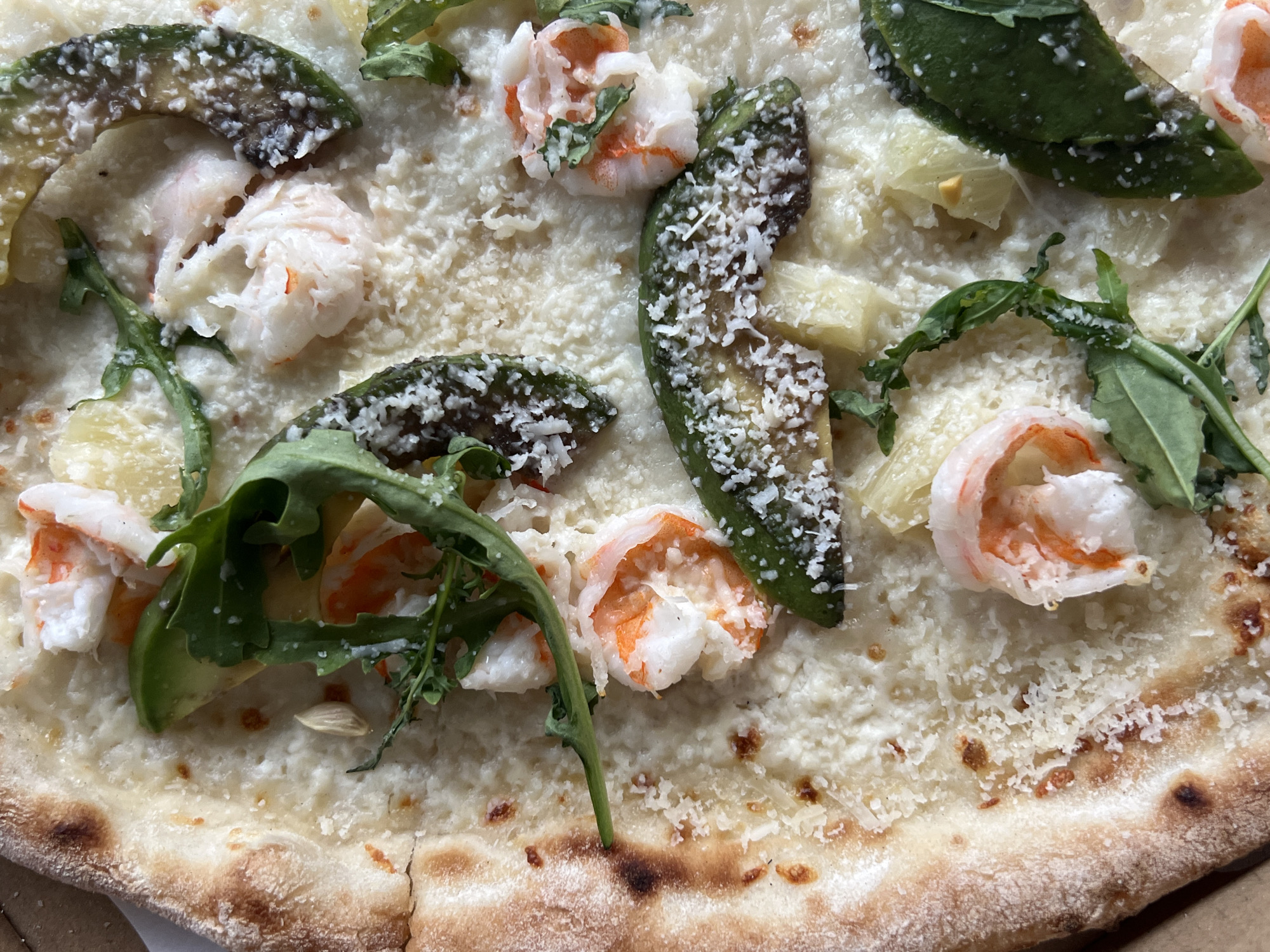A close-up, overhead shot of a pizza topped with white sauce, shrimp, avocado, and arugula. The pizza crust is golden brown with some darker, charred spots. Shredded Parmesan cheese is sprinkled over the toppings. The image is detailed, showcasing the textures and colors of the ingredients. The pizza is cut into wedges. There are no people or any other objects visible in the frame. The scene is presented on a flat surface. This image does not depict any location in the city of Briukhovychi, Ukraine.