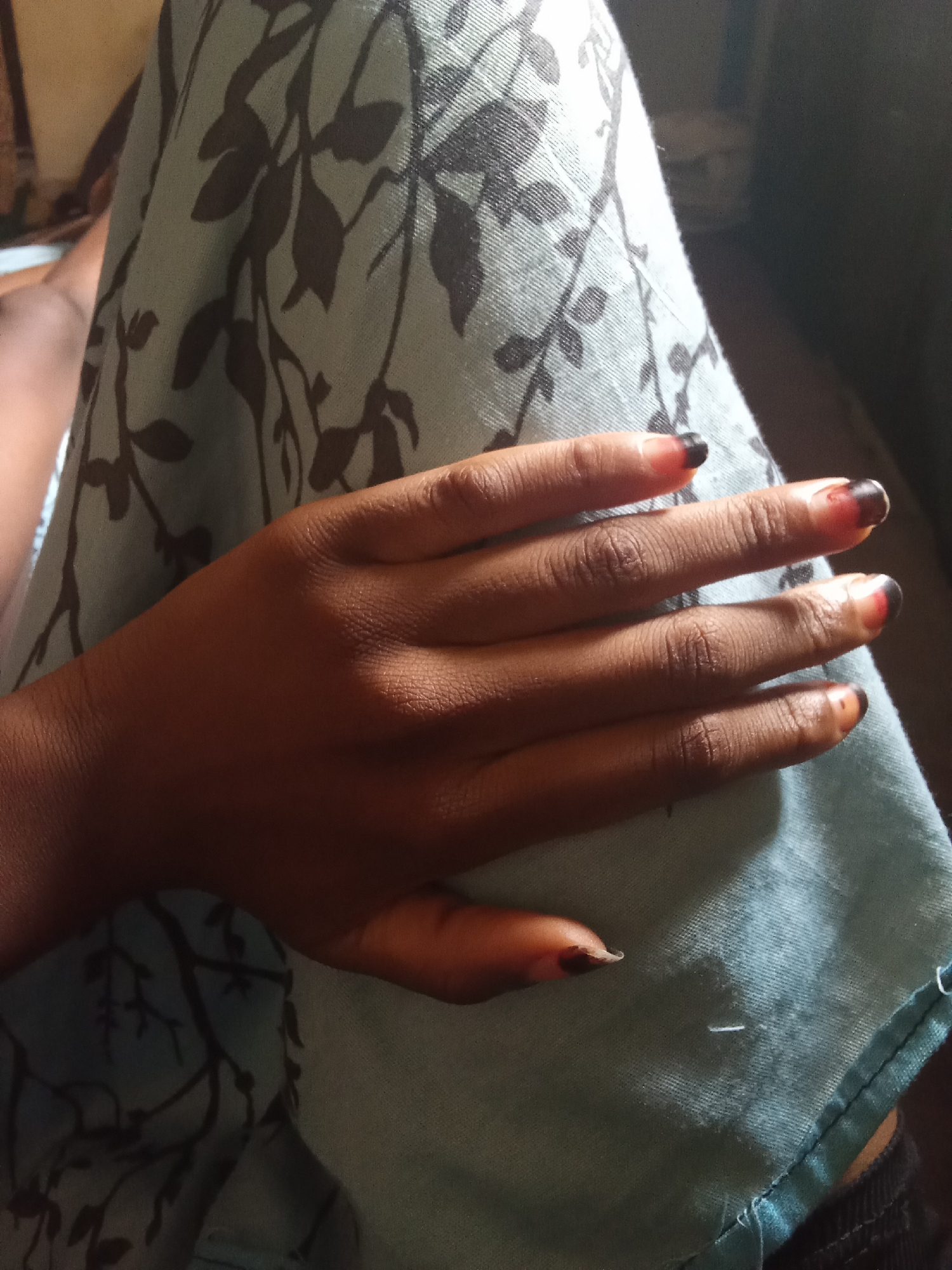 The image shows a close-up of a person's hand resting on a light blue fabric with a dark floral pattern. The hand is a medium brown skin tone, and the fingernails are painted with a dark color, possibly black, on the tips and a reddish-orange near the cuticle. The fabric appears to be a garment or upholstery, with visible seams and stitching along the bottom right edge. The lighting suggests an indoor setting, possibly daytime due to the soft shadows and even illumination. There is no visible text in the image. The overall impression is one of a casual, personal moment captured in a still photograph.