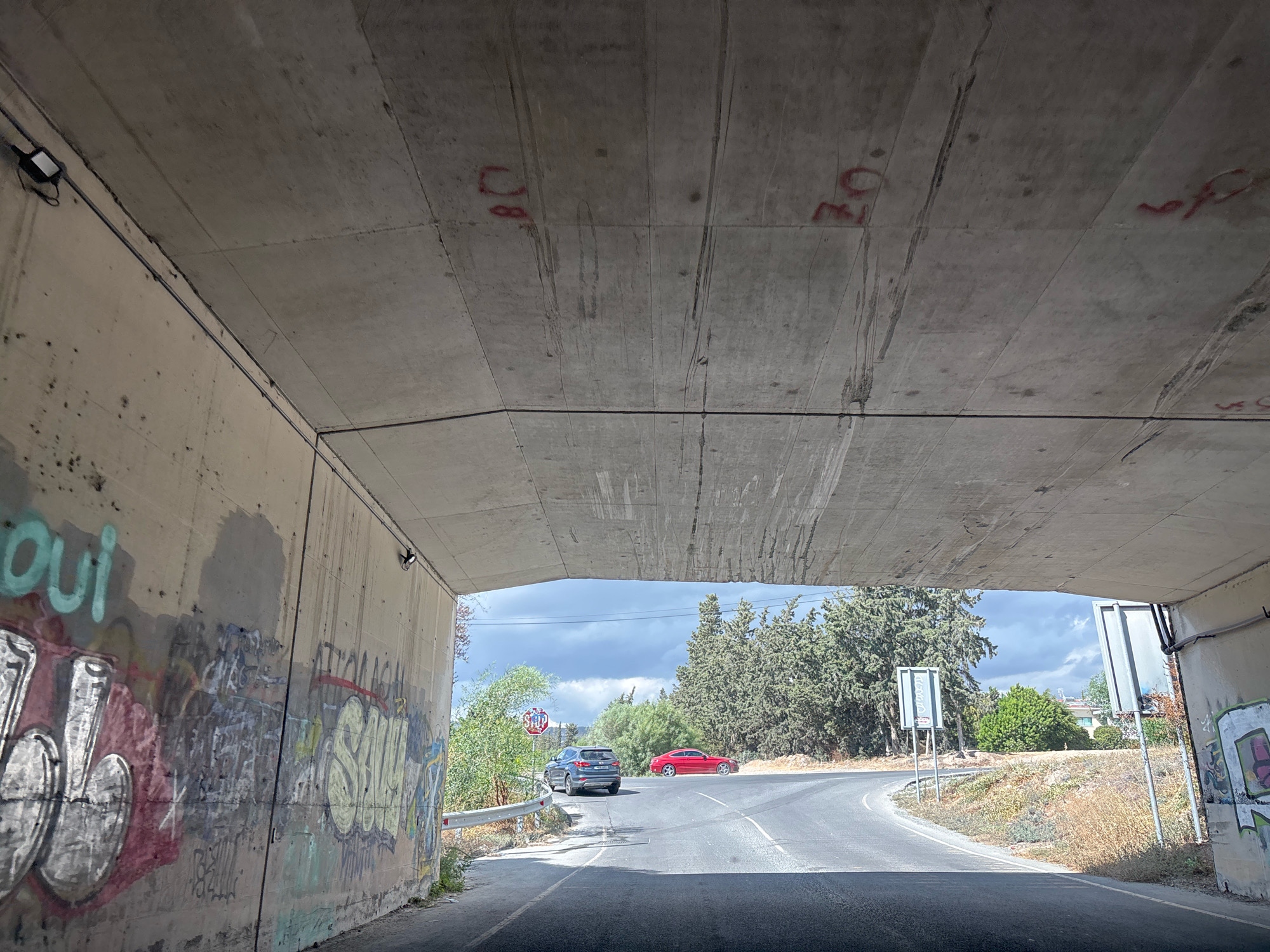 The image shows a view from under a concrete overpass in Agios Athanasios Municipality, Cyprus. The overpass has graffiti on its interior walls. A road curves out from under the overpass, with trees and vegetation lining its sides. A red sports car and a dark SUV are visible on the road. A stop sign is present near the SUV. The sky is cloudy.