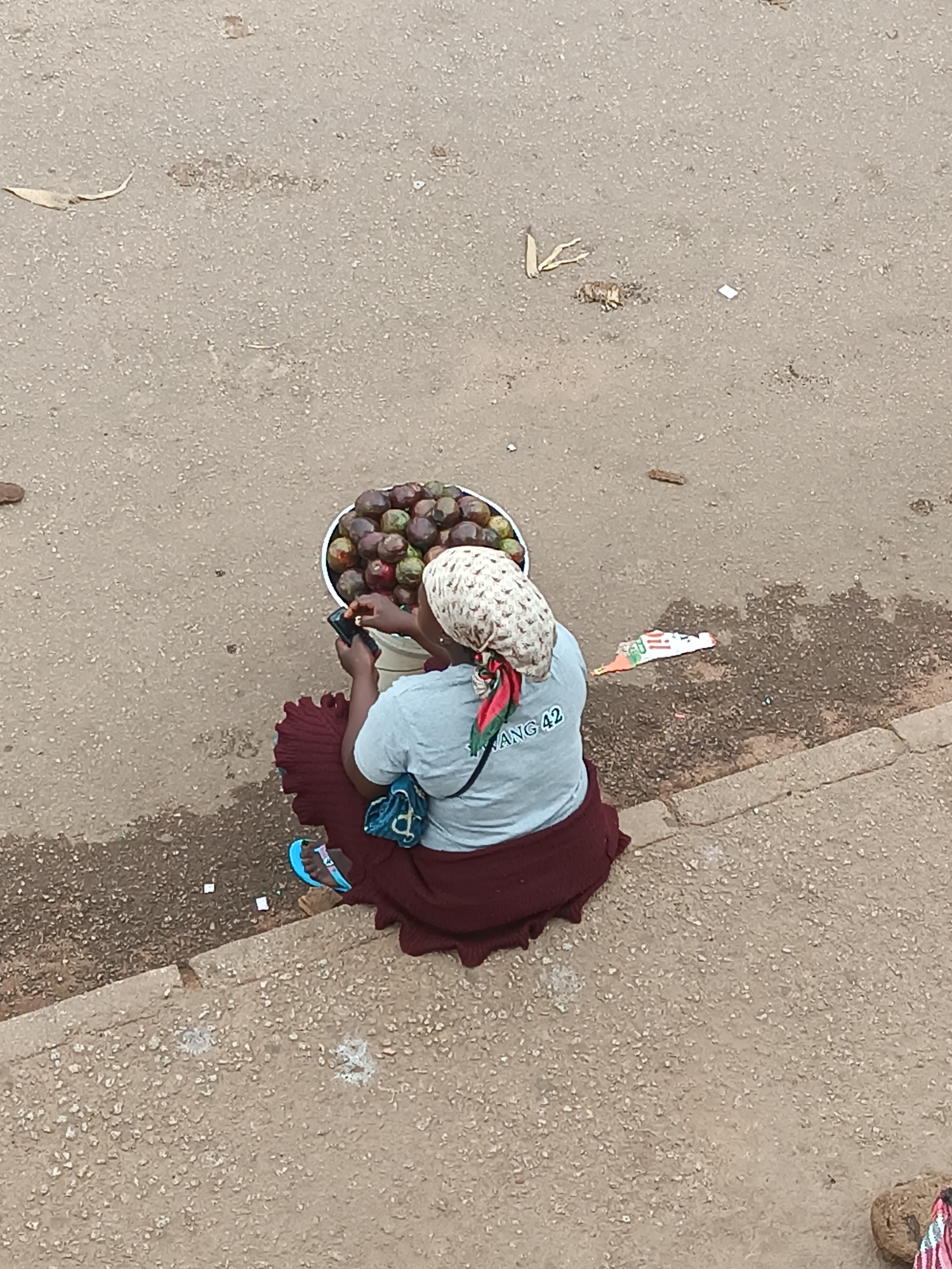 An overhead shot captures a person, likely a woman, sitting on the ground next to a large bucket of fruit. The individual is seen from behind, wearing a patterned cream headscarf, a light blue t-shirt, and a dark red skirt or wrapped fabric. A blue bag is slung over their shoulder, and a light blue flip-flop is visible on one foot.
The woman holds a black smartphone in her hands, looking down at the screen, indicating she is actively using the device. In front of her is a white plastic bucket brimming with numerous small, round fruits that range in color from green to reddish-purple, resembling plums or similar stone fruits.
The setting is an outdoor, unpaved or lightly paved area, possibly a roadside or marketplace in Bukuru, Nigeria. The ground is a mix of light-colored concrete or compacted dirt with darker, damp-looking patches and scattered debris such as dry leaves and small pieces of litter. A raised curb defines the edge of the immediate sitting area. The bright lighting suggests it is daytime. Visible text on the back of the woman's t-shirt reads "ANG 42".