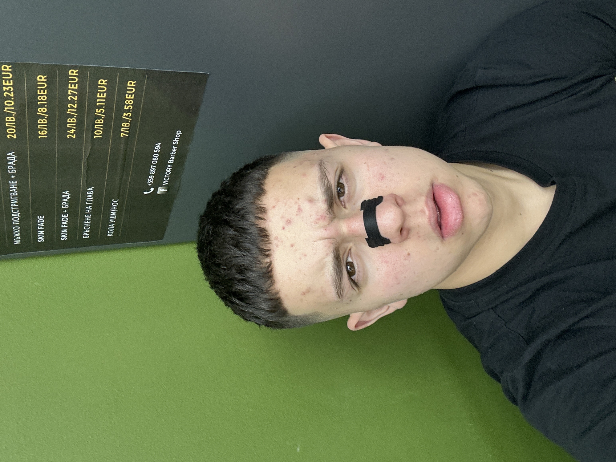 A young man with a black nose strip is posing for a selfie indoors, with a green wall visible in the background. A price list for "VICTORY Barber Shop" is partially visible, listing services in Bulgarian and their prices in both Bulgarian Leva (LV) and Euros (EUR). The prices are: "МЪЖКО ПОДСТРИГВАНЕ + БРАДА" (Men's haircut + beard) - 20ЛВ./10.23EUR, "SKIN FADE" - 16ЛВ./8.18EUR, "SKIN FADE + БРАДА" (Skin Fade + Beard) - 24ЛВ./12.27EUR, "БРЪСНЕНЕ НА ГЛАВА" (Head shave) - 10ЛВ./5.11EUR, "КОЛА УШИ/НОС" (Ear/Nose wax) - 7ЛВ./3.58EUR. The shop's phone number is listed as +359 897 080 594. It is presumed to be in Zlatograd, Bulgaria, based on the language and currency.
