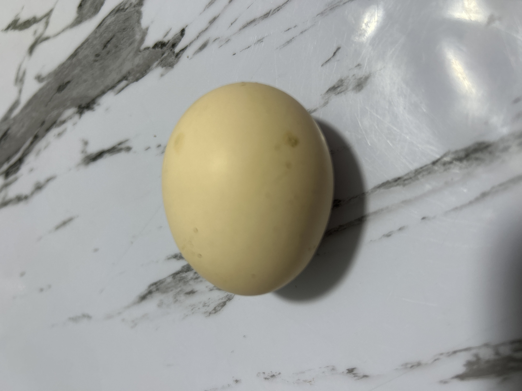 A single, light yellow egg rests on a marble-patterned surface. The surface is predominantly white with dark gray, vein-like markings. The egg is positioned slightly to the left of the center of the frame, casting a soft shadow to its left and below. The lighting appears to be from the top left, creating highlights on the egg's surface. The scene is static and does not depict any specific location within Дедемель, Kyrgyzstan, as there are no identifying features of that city present in the image.