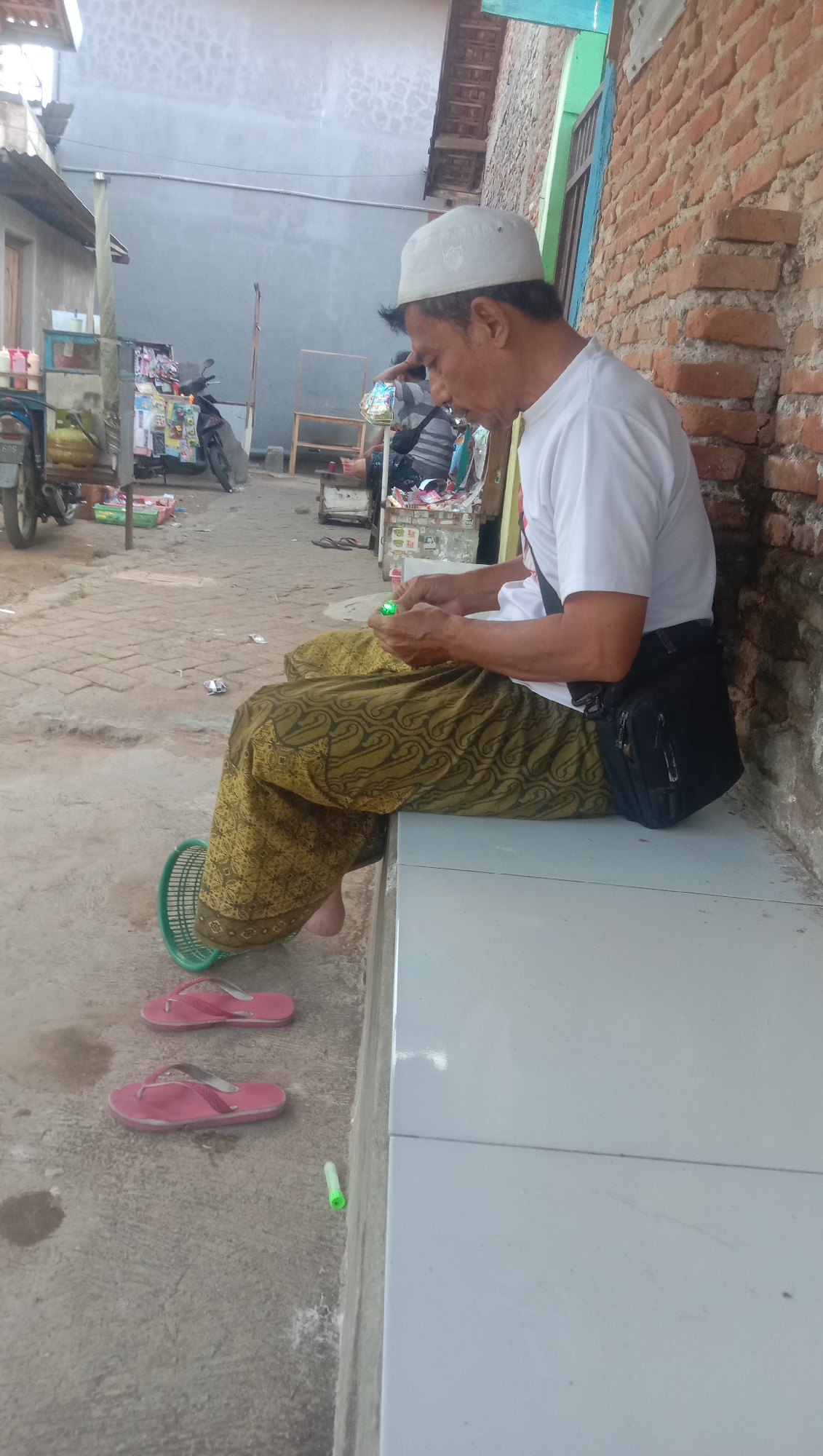 In Karang Anyar, Indonesia, a man wearing a white skullcap and a white t-shirt is sitting on a concrete bench. He is also wearing a patterned sarong. In his hands, he is holding a small green object, possibly a toy or a light. A black shoulder bag is slung across his chest. In front of him on the ground, there are two pink flip-flops and a green object, which appears to be a pen or a small marker. To the left of the man, a green plastic basket is visible. The background shows a brick wall and a street with some market stalls and a motorcycle. The overall scene depicts a casual moment in a semi-urban environment.