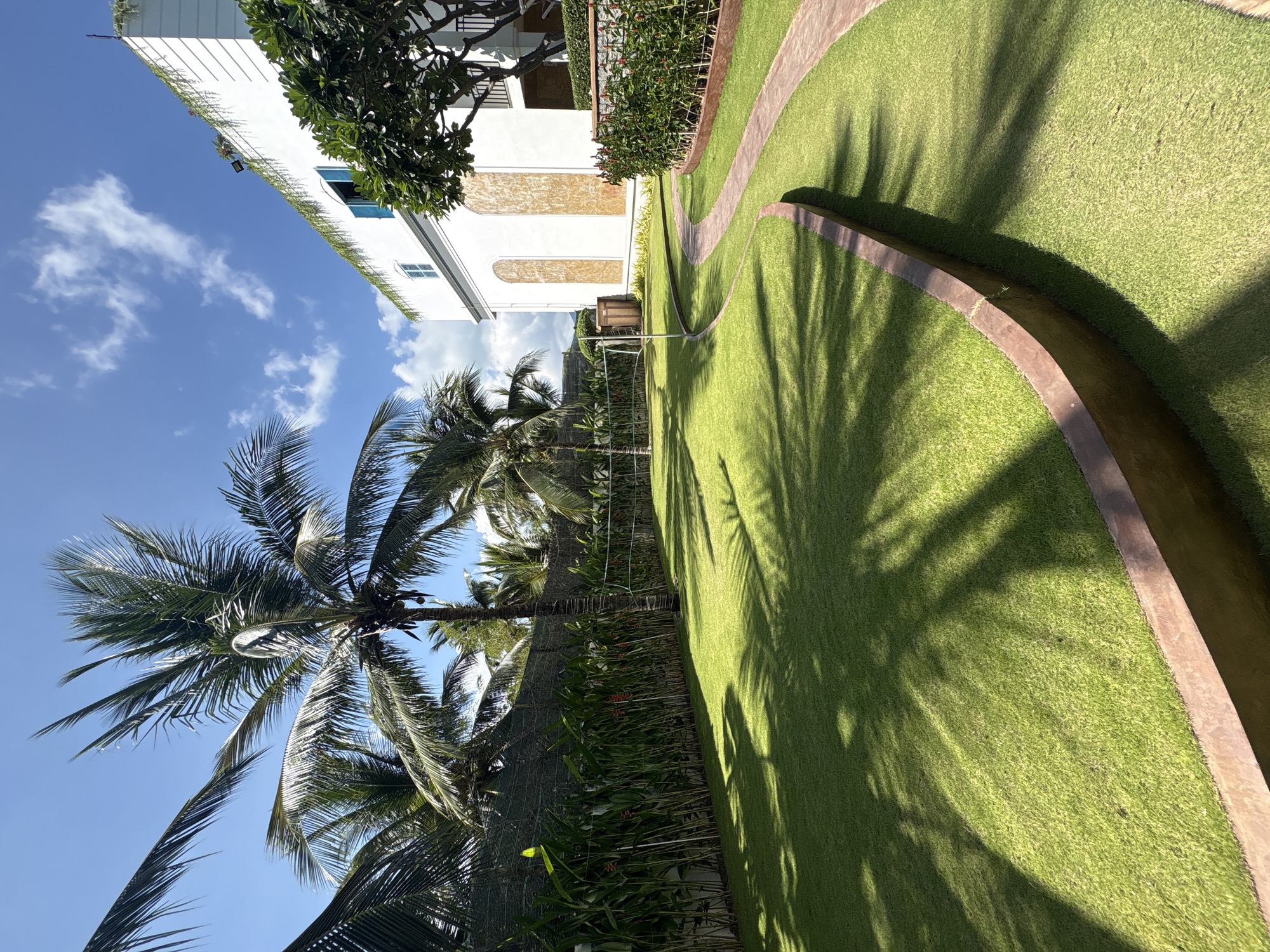 The image shows a tropical scene in Bac Nha Trang Ward, Vietnam. A white building with architectural details is visible on the left. Palm trees are in the foreground against a blue sky with scattered clouds. A green lawn extends across the middle ground, bisected by a winding, brown-edged water feature or path. Shadows of palm trees fall across the lawn. Lush vegetation and fencing line the perimeter of the lawn. No people are visible in the image.