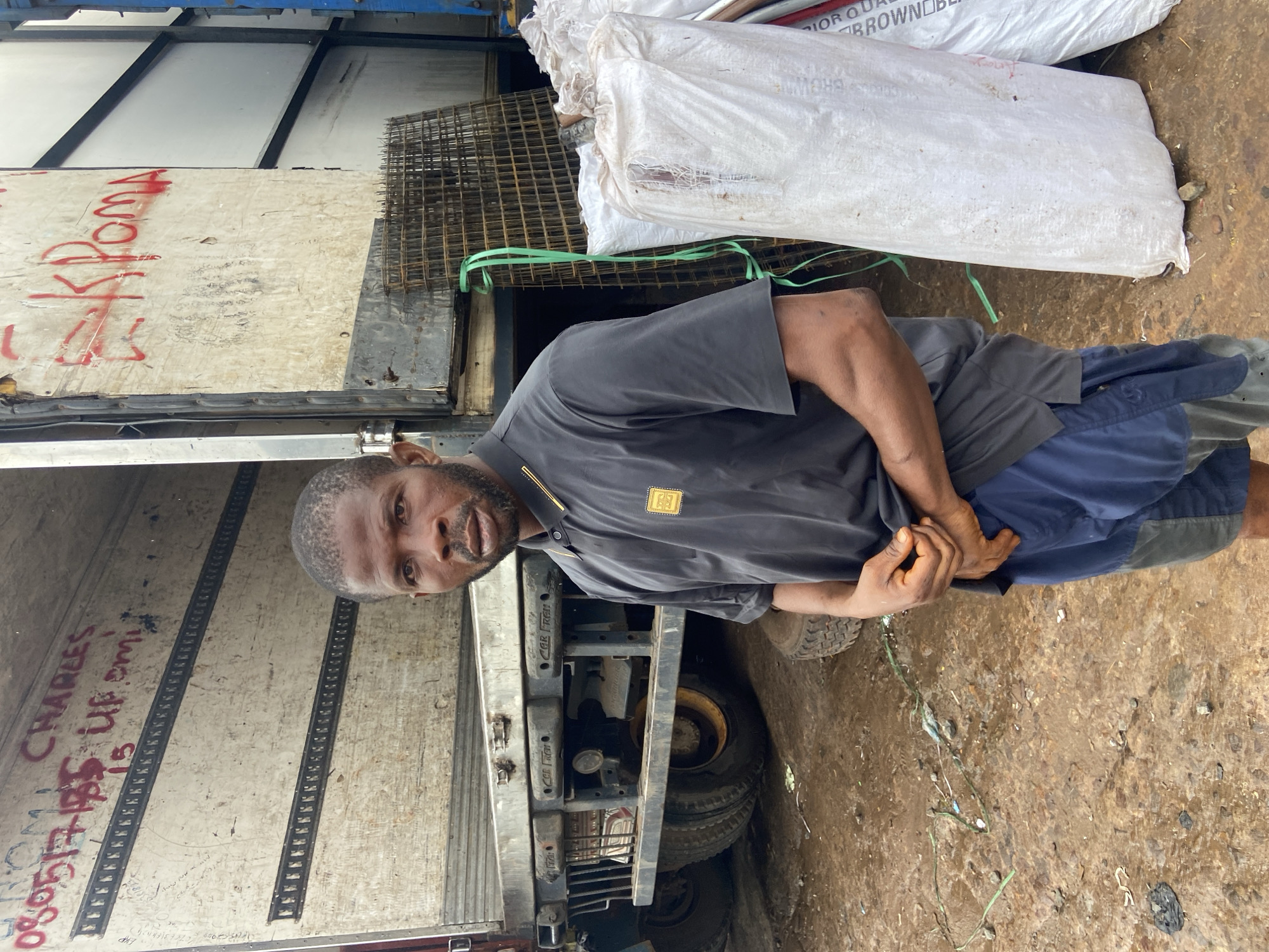 This media file depicts a man standing next to a truck in Ogbunike, Nigeria. The man is wearing a dark gray polo shirt and blue shorts. He has his hands clasped in front of him. Behind the man, the truck is loaded with what appears to be sacks of material, possibly fertilizer or feed, and some metal mesh. Red graffiti is visible on the side of the truck, including the word "ROM" and what looks like a phone number. Further text in red on the truck reads "CHARLES" and "UP EMI". A part of another truck is visible to the left. The ground is a mixture of dirt and debris. The lighting suggests it is daytime. The overall scene appears to be an industrial or commercial area, likely a transport hub or storage yard, given the presence of the trucks and stored materials. The man's posture and expression are neutral, with no discernible strong emotions.