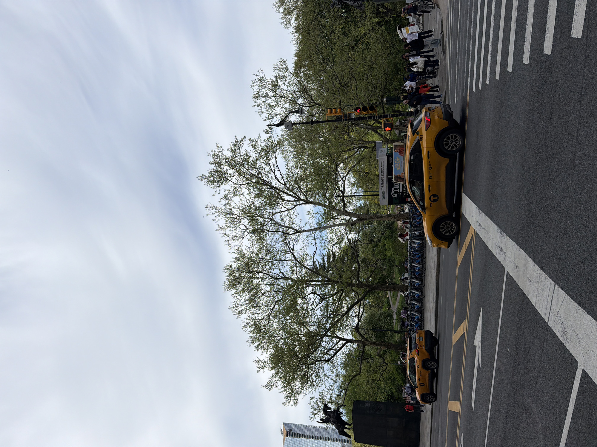 The image captures a New York City street scene, likely in the vicinity of Central Park. Two yellow taxis are positioned on the right side of the frame, near a pedestrian crosswalk where a group of people are waiting to cross the street. A row of bicycles is stationed along the sidewalk to the left. The scene is partially obscured by trees. In the distance, there is a partially obstructed view of a monument featuring a statue of a person on horseback. The sky is overcast with varied cloud formations. A modern skyscraper is visible in the background.
