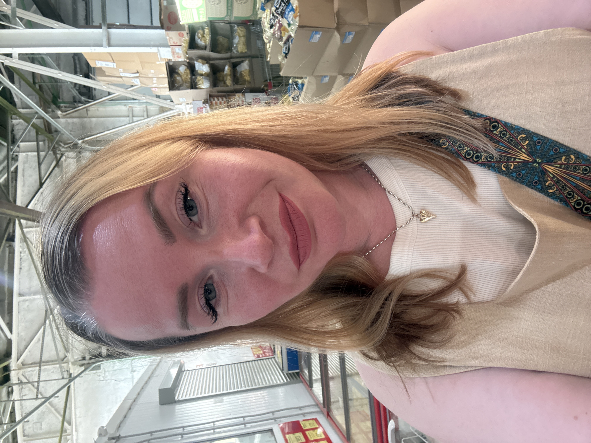 A forensic selfie captured in a commercial environment, likely a wholesale store or warehouse, in Pankovka, Russia. The image features an adult female with fair skin and long, light blonde/brown hair, displaying visible grey regrowth at the roots along her part and forehead. She has light eye makeup, including dark mascara on her upper lashes, and light pink/mauve lipstick. Her facial expression displays a faint, closed-mouth smile. She is wearing a light beige or cream ribbed tank top layered under a sleeveless, V-neck garment of a similar color and linen-like texture. A silver-colored chain necklace with a small, gold-toned, pointed pendant rests on her chest. A decorative, patterned strap, featuring blue, gold, red, and black geometric motifs, crosses diagonally from her left shoulder area.
The background behind her indicates an indoor, industrial-style building with high ceilings, exposed white metal trusses, beams, and conduits. To her left, white corrugated metal surfaces, possibly roll-down doors or refrigeration units, are visible, along with red and white signage displaying numbers like "363" and "175." To her right, stacks of cardboard boxes containing various packaged goods, some in clear plastic bags, are arranged on shelves and stacked on the floor. The overall lighting is bright and even, characteristic of a commercial interior.