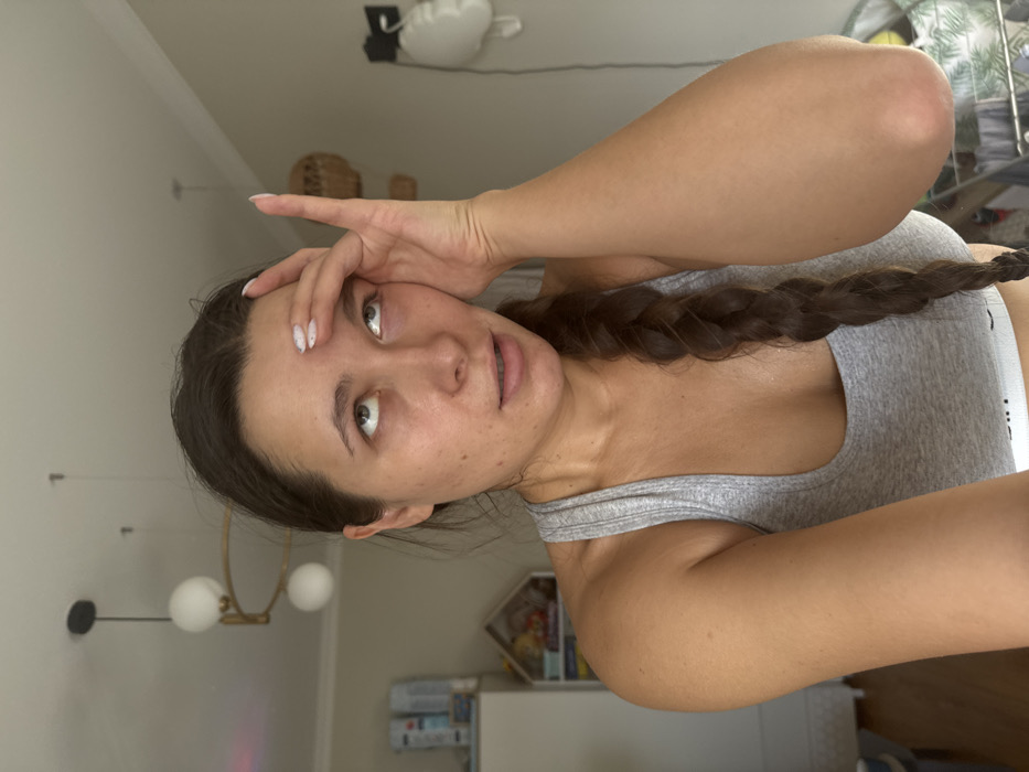A female individual, appearing to be a young adult, is depicted in a close-up, chest-up shot indoors in the city of Селекционер-2 Снт, Russia. She has dark hair styled in a long braid resting on her right shoulder and chest, and is wearing a light grey athletic crop top. Her eyes are rolled upwards, and her left hand is placed on her forehead with fingers splayed, partially obscuring her left temple and upper forehead. Her mouth is slightly open. The background reveals a light-colored interior room with a white ceiling featuring two modern pendant light fixtures: one with two spherical white globes on a horizontal gold-toned bar, and another single spherical globe fixture. To the subject's left, a white cabinet, a small wooden house-shaped decorative shelf containing items, and a portion of a window are visible. To the subject's right, a woven decorative wall hanging, a white wall-mounted sculptural mask, and a partial view of a clothes drying rack are present.