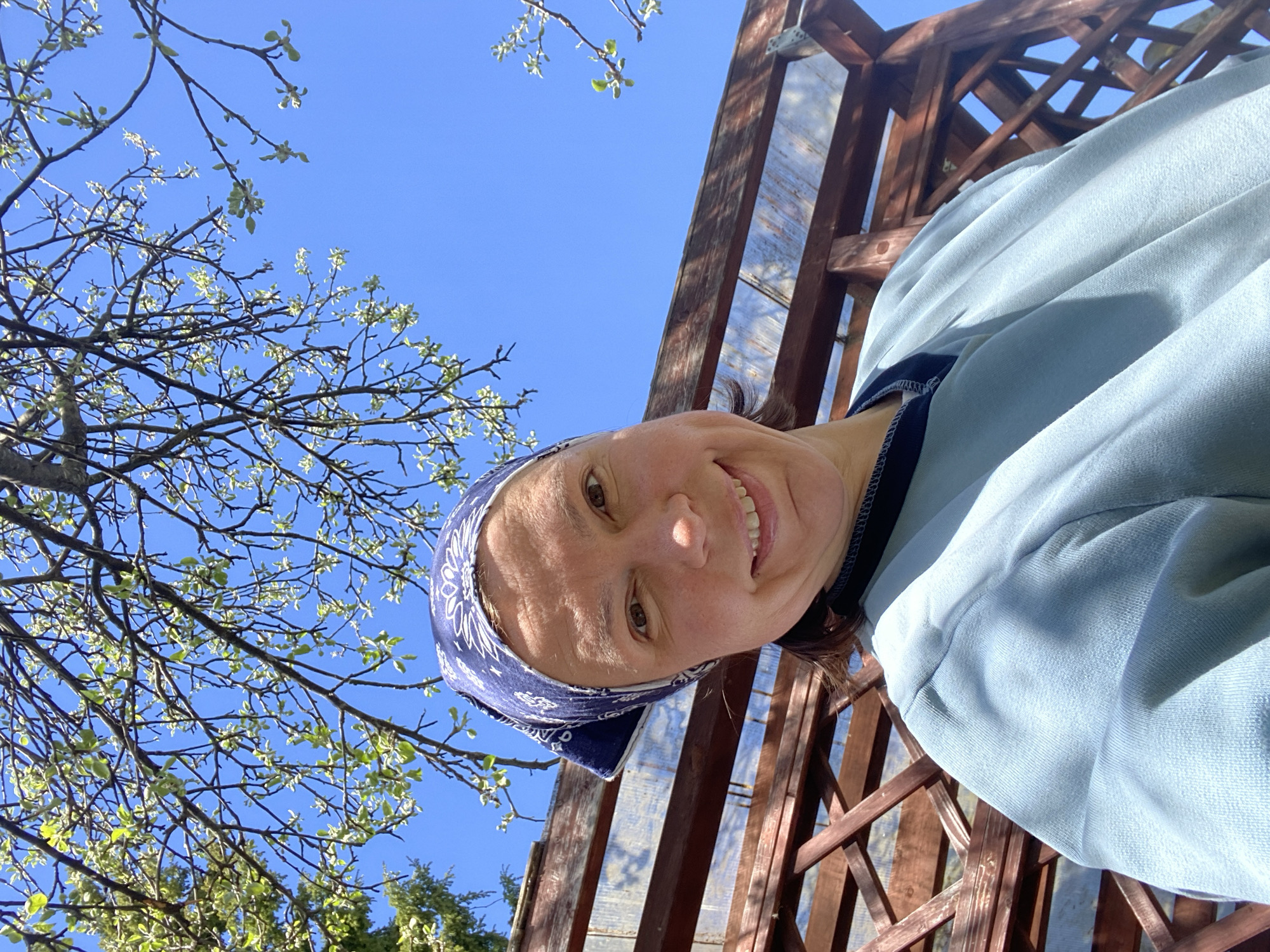 A person with a bandana is smiling at the camera, positioned underneath a wooden lattice structure with foliage and branches visible against a blue sky. The person is wearing a light blue garment with a dark blue trim. The location is not discernible as being in the city of Прикамский, Russia, based on the image alone.