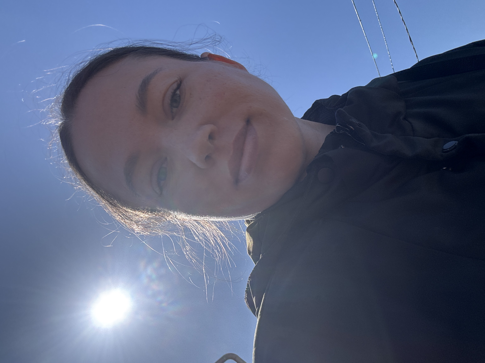 A low-angle selfie of a woman with fair skin and dark hair pulled back, taken outdoors on a sunny day. The sky is bright blue. The woman is wearing a dark-colored jacket. Electrical wires are visible in the upper right corner. The sun is shining brightly in the lower left corner. The picture was taken in Ферма, Russia.