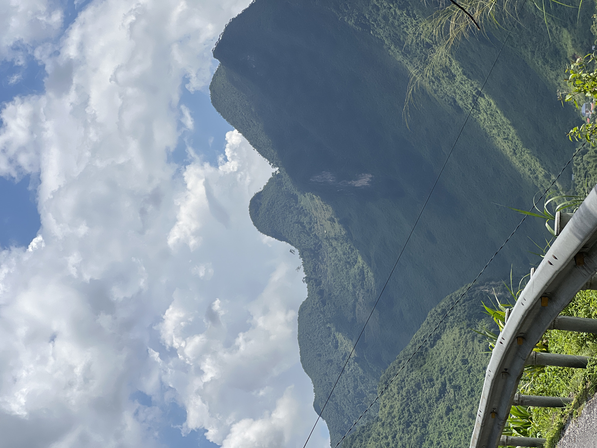 A mountainous landscape in Lùng Tám Commune, Vietnam, is captured under a cloudy sky. The foreground features a guardrail along a road, with lush green vegetation growing alongside. Beyond the guardrail, steep, verdant mountains dominate the scene, their slopes covered in dense foliage. A few power lines stretch across the frame, diagonally crossing the mountains. The lighting suggests it is daytime, with shadows indicating the presence of clouds. The image is a natural, unposed photograph of the rural, mountainous terrain.