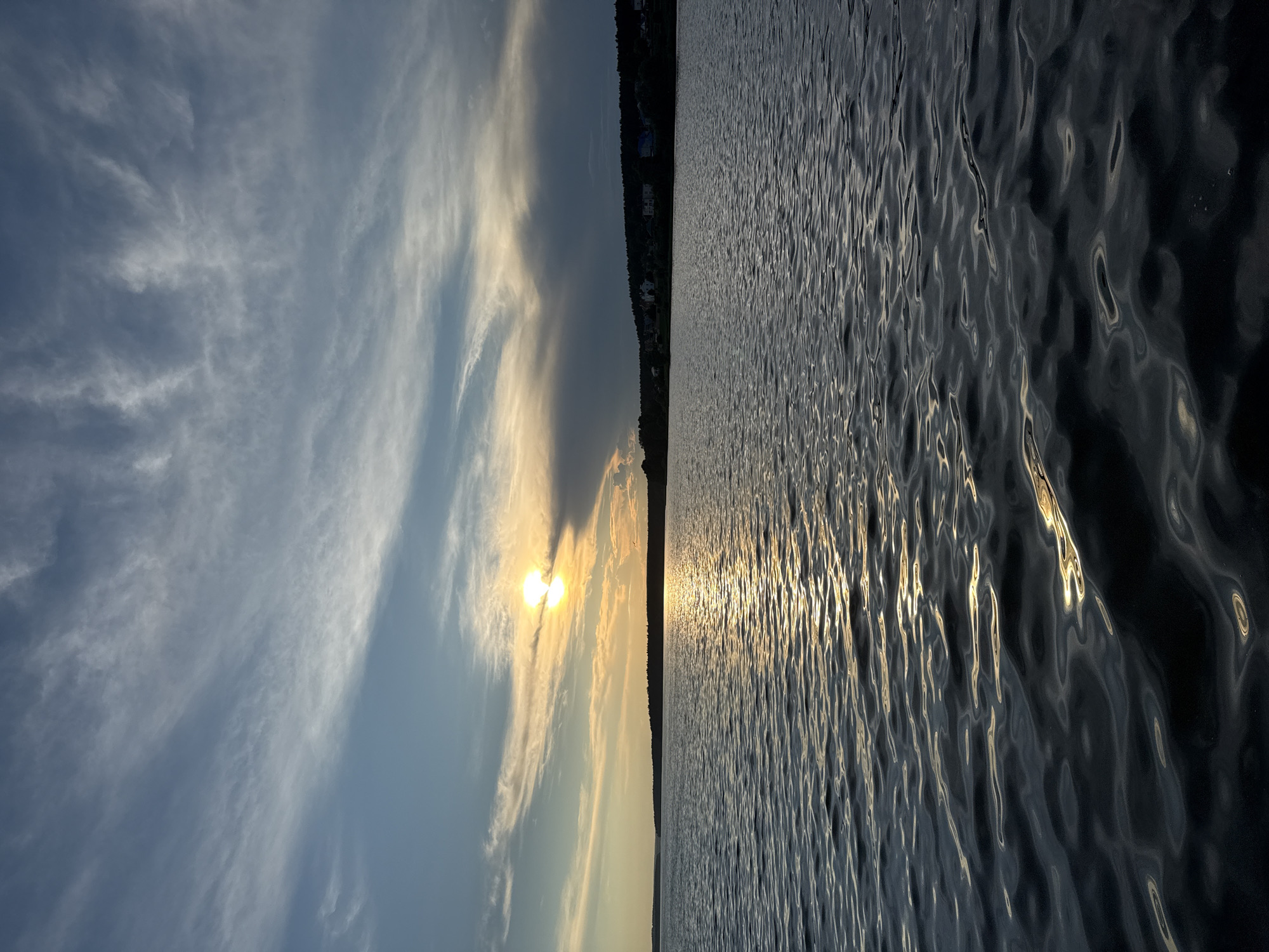 The image captures a serene sunset over the Kama River in Votkinsk, Russia. The sun, a bright orb partially obscured by wispy clouds, casts a warm golden glow across the sky and its reflection shimmers on the rippling water. In the distance, a tree-lined shore is dotted with houses, suggesting a settlement or village. The sky is a gradient of blues, transitioning from a light, hazy blue near the horizon to a deeper blue overhead. The water's surface is a deep, dark blue, disturbed by gentle waves that catch the light, creating streaks of gold and silver. The overall scene is tranquil and atmospheric, evoking a sense of peace and natural beauty at the end of the day.