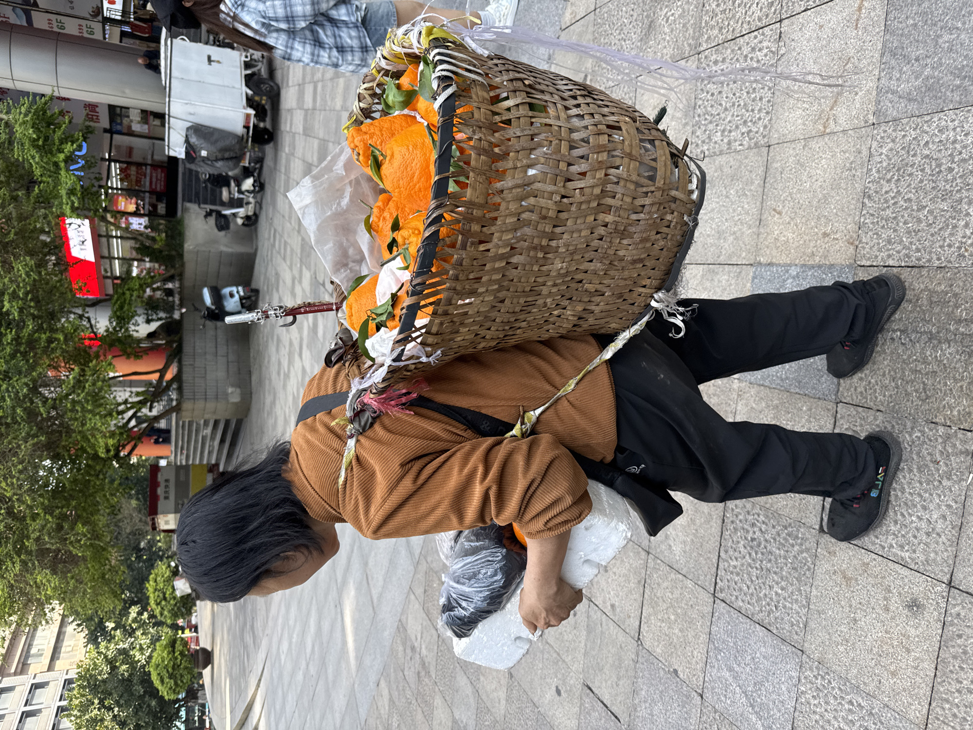 A woman is walking on a gray brick sidewalk in Yuzhong District, China. She has a large woven basket on her back filled with oranges with leaves. The basket is supported by a rope or strap that goes over her shoulders. She wears a brown corduroy jacket, black pants, and black shoes. She is carrying something white in her right hand. In the background, there are buildings, trees, and other people.