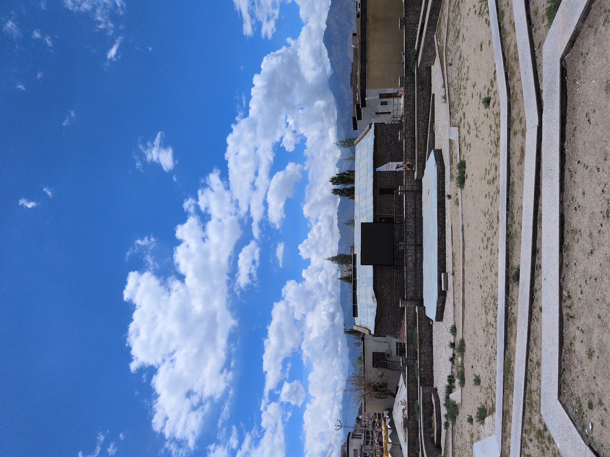 The photograph captures a sunlit outdoor space in Leh, India, likely during the day. The foreground features a landscaped area with sand and sparse vegetation, bordered by concrete pathways and retaining walls. In the mid-ground, there's a raised, light-colored platform that appears to be a stage or seating area. Behind this, a low-slung building with a corrugated metal roof and stone walls is visible. A large, dark rectangular screen is mounted on the building. Several trees stand behind the building, and in the background, mountains with snow-capped peaks rise against a vibrant blue sky dotted with fluffy white clouds. To the left of the main building, there are glimpses of other structures and utility poles, suggesting an urban or semi-urban environment. A banner with text and an image is visible near the building's entrance, hinting at a cultural or informational display. The scene is devoid of people.