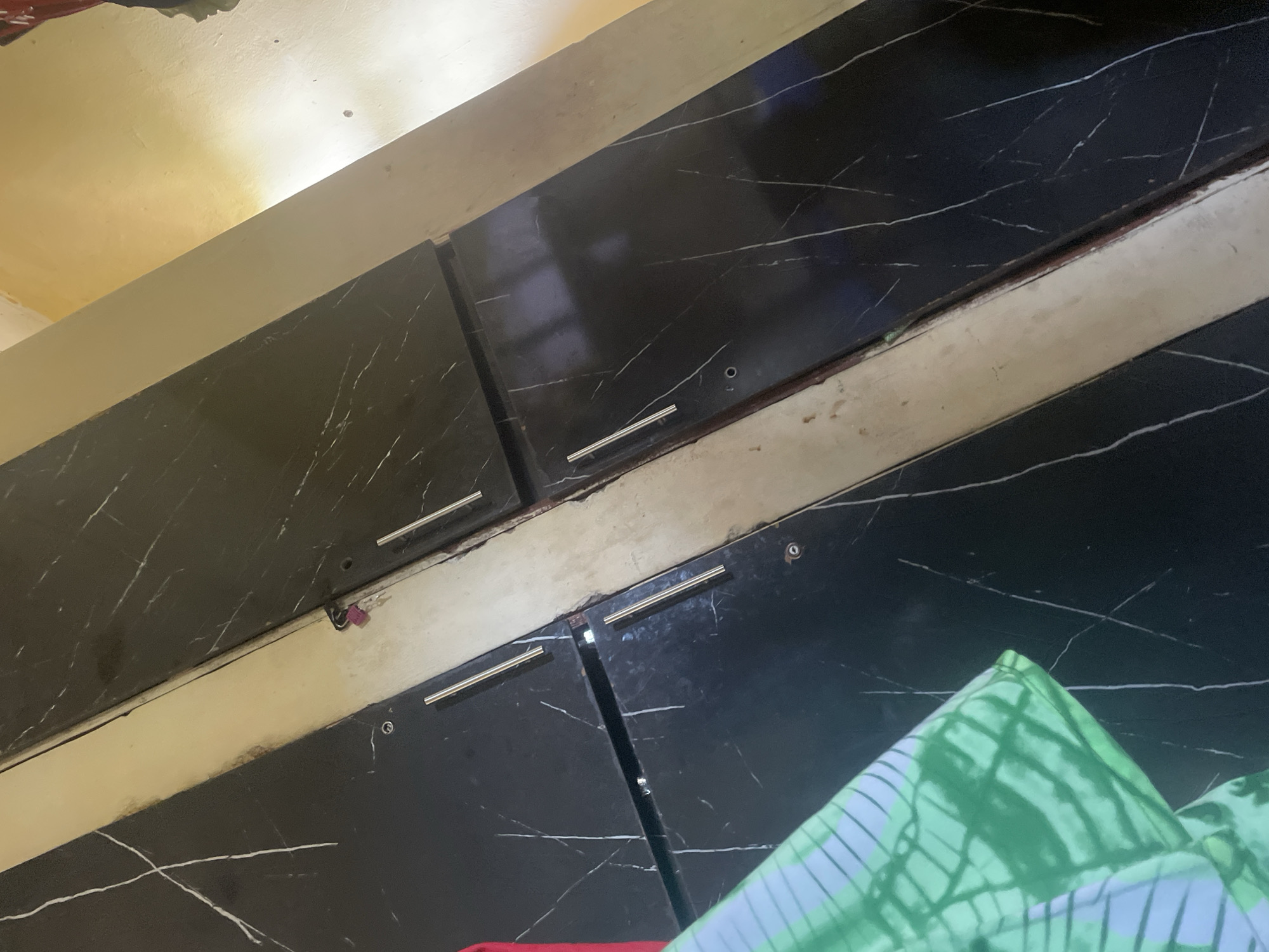An indoor scene in Jalingo, Nigeria, shows a close-up view of black marble-patterned cabinets with silver handles. The cabinets appear to be part of a wardrobe or a similar furniture piece. A portion of a green and white patterned fabric is visible in the lower left corner of the image. The lighting suggests an interior space with ambient light coming from the right side, casting shadows and highlighting the textures of the cabinets and the wall. The overall impression is one of a residential or commercial interior.