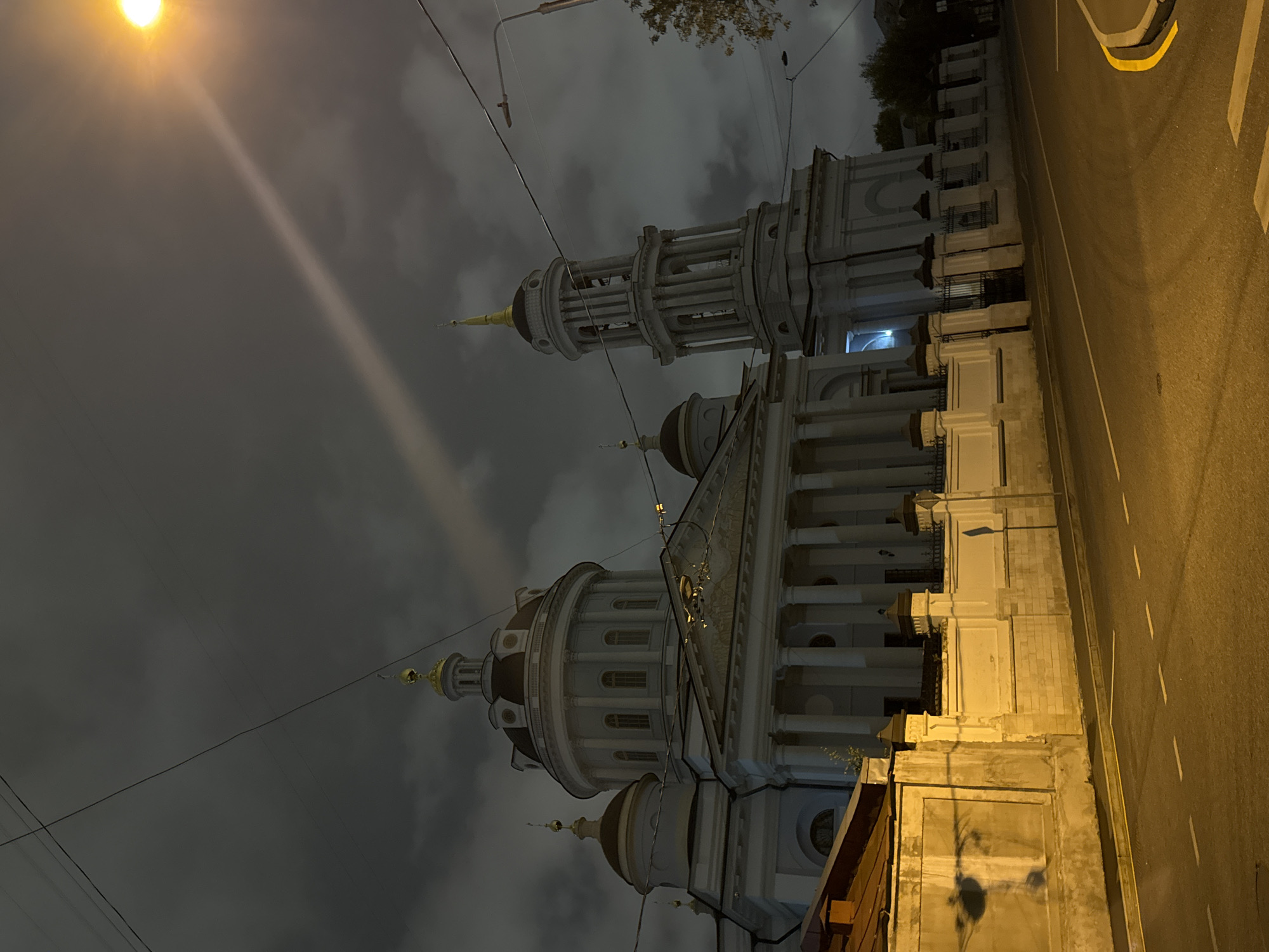 The image depicts a nighttime view of the Church of the Ascension of the Holy Cross (Khram Vozneseniya Khristova) in Moscow, Russia. The church, a prominent architectural landmark, is illuminated, with its white facade and golden domes standing out against a dark, cloudy sky. A tall bell tower is visible to the right of the main structure. The foreground shows a street with asphalt paving and yellow lane markings, suggesting it's a road with traffic. A sidewalk or wall runs along the side of the church, separating it from the street. Overhead power lines are visible, crossing the sky. A street lamp casts a bright light and a beam across the cloudy sky. No people are visible in the image. The overall impression is one of a historical and religious site at night, with typical urban infrastructure present.