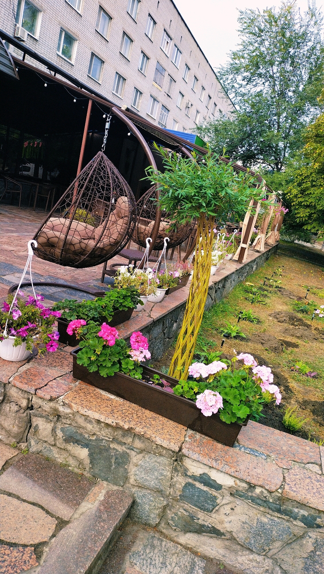 The image shows an outdoor seating area in front of a multi-story brick building, likely a cafe or restaurant, located in Horishni Plavni, Ukraine. The foreground features a stone staircase leading up to a stone wall, on top of which are several planters filled with pink and purple flowers. Behind the planters, two large, woven hanging chairs are visible, suspended from a metal frame. The chairs are equipped with plush brown cushions. In the background, the brick building has numerous windows and an air conditioning unit on the left side. A large tree with green foliage stands to the right of the building. The ground beyond the seating area appears to be a garden with tilled soil and some small plants. The overall scene suggests a pleasant outdoor space designed for relaxation.