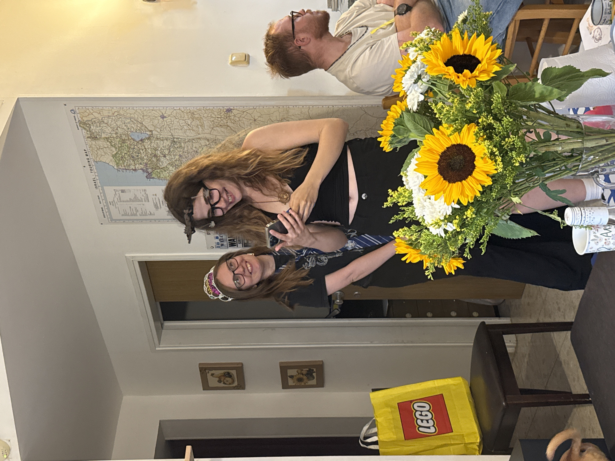 The image depicts an indoor scene, potentially in a residence within the city of Modiin-Maccabim-Reut, Israel, suggested by a map of Israel hanging on the wall. Three people are visible: two women who seem to be taking a selfie and a man in the background. The women are positioned in the center, one wearing glasses and a tiara, the other wearing glasses. The man is looking upwards. A bouquet of sunflowers and white flowers is prominent in the foreground. Additional objects include a yellow LEGO bag, framed pictures on the wall, a chair, and a cup.