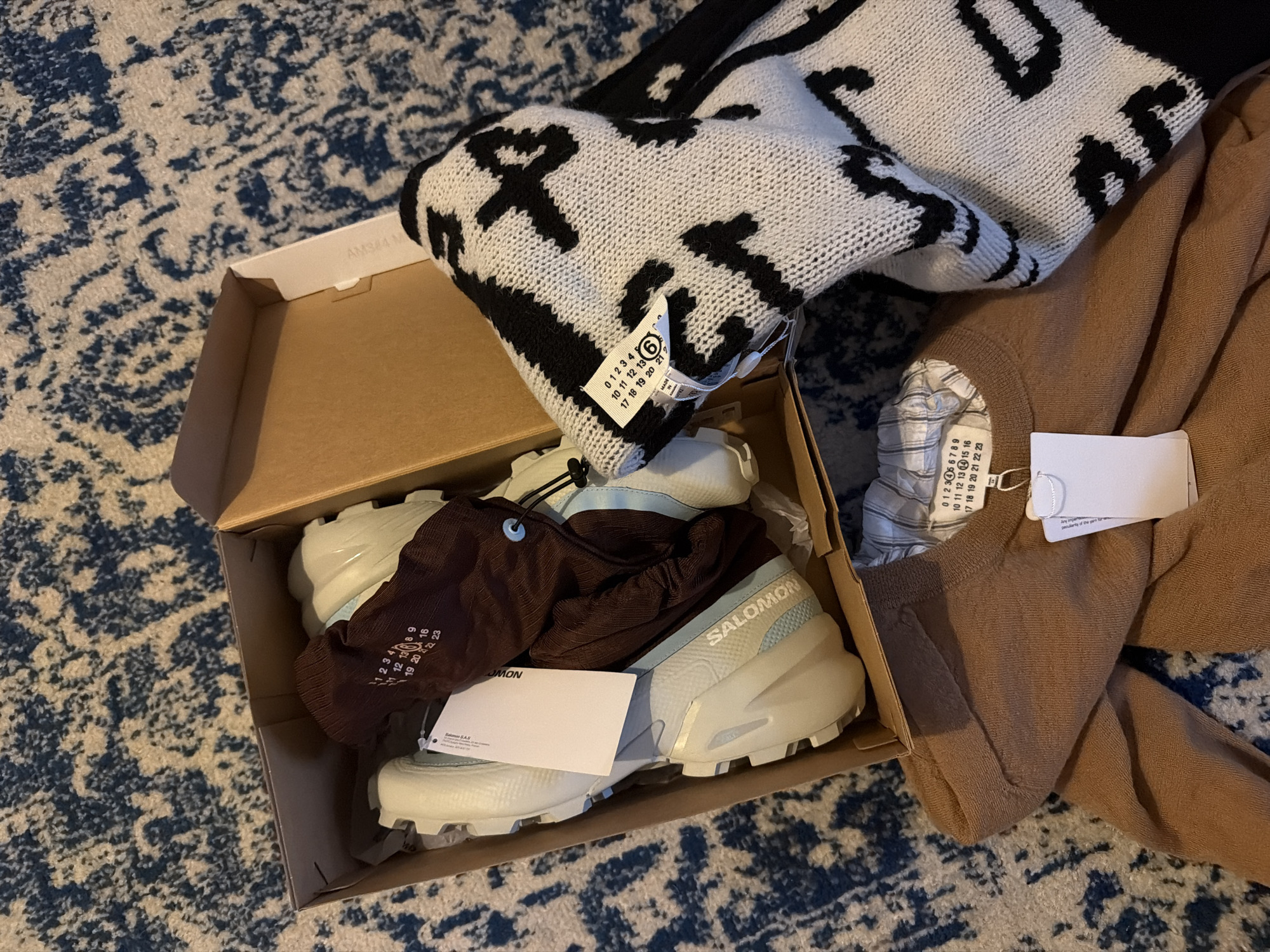 The image shows a cardboard box filled with items placed on a rug with a blue and white patterned design. Inside the box, there are light-colored Salomon sneakers, covered partially by a brown drawstring bag. A white card is placed on top of the bag. Next to the box, there is a pile of clothing consisting of a black and white patterned knit sweater and a tan-colored sweater. Each sweater has a white tag attached.