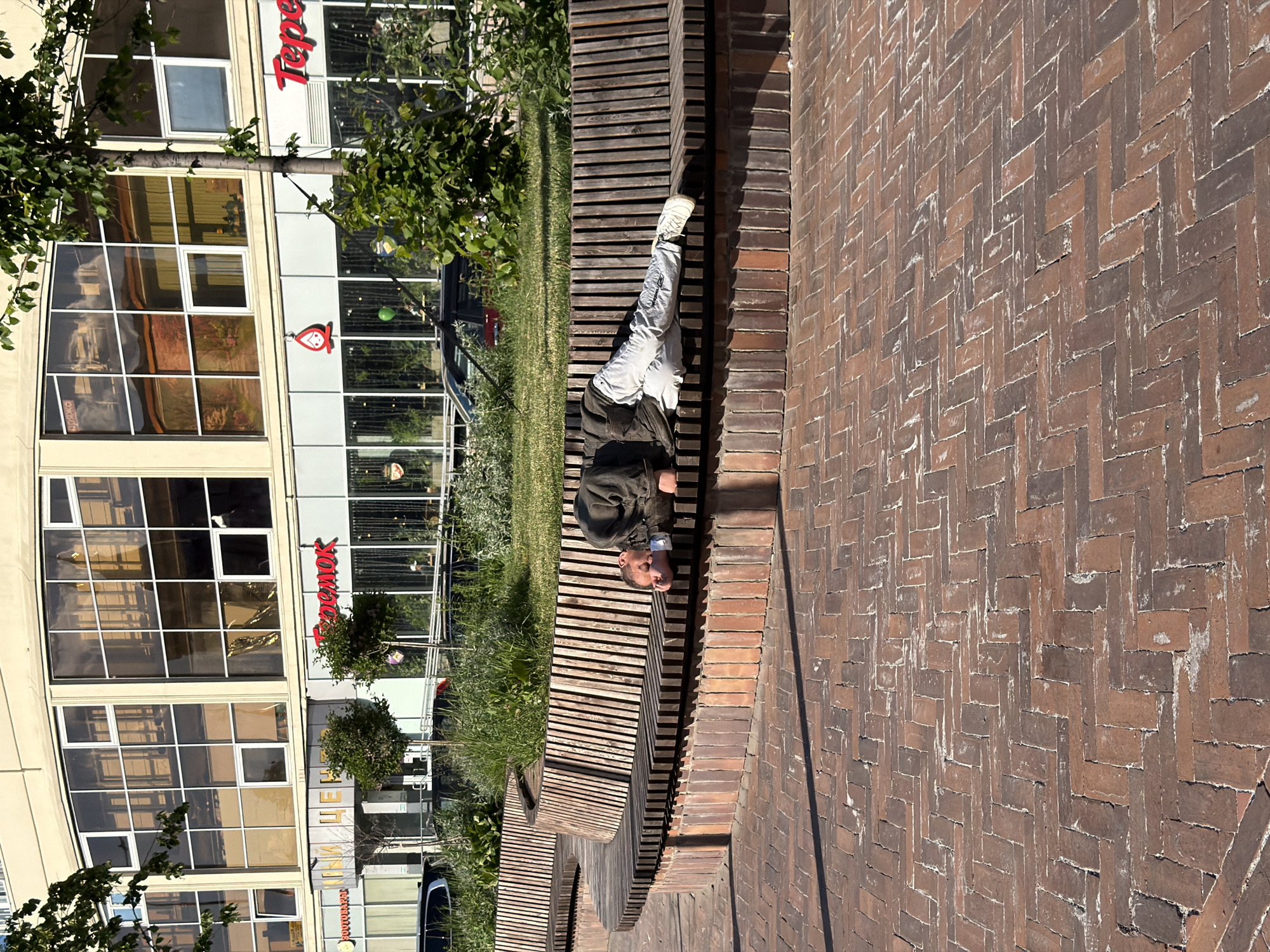 An outdoor, sunlit scene captures a man resting on a wooden slatted bench. He is lying on his back, with his head positioned toward the left of the frame (if the image were rotated upright) and his feet extended to the right. He is wearing light-colored trousers, white sneakers, and a dark jacket. The bench has a distinctive wavy, serpentine design and is set amidst a patch of green grass and small trees.
Behind the man and the grassy area are modern buildings with large, reflective glass windows, suggesting commercial or office spaces. Reflections in the windows show parts of the surroundings, including trees and possibly vehicles. To the right of the bench, a patterned brick walkway or wall is visible, made of reddish-brown bricks laid in a herringbone pattern. The bright sunshine casts distinct shadows, indicating it is daytime.
Visible text on one of the buildings includes "Tepe" (part of a sign) and "Теремок" (Teremok), identifiable as a Russian fast-casual restaurant chain, along with its distinct red logo resembling a simplified figure. This detail aligns with the given location context of Moscow, Russia. The overall atmosphere is tranquil, with the man seemingly asleep or deeply relaxed on the bench.