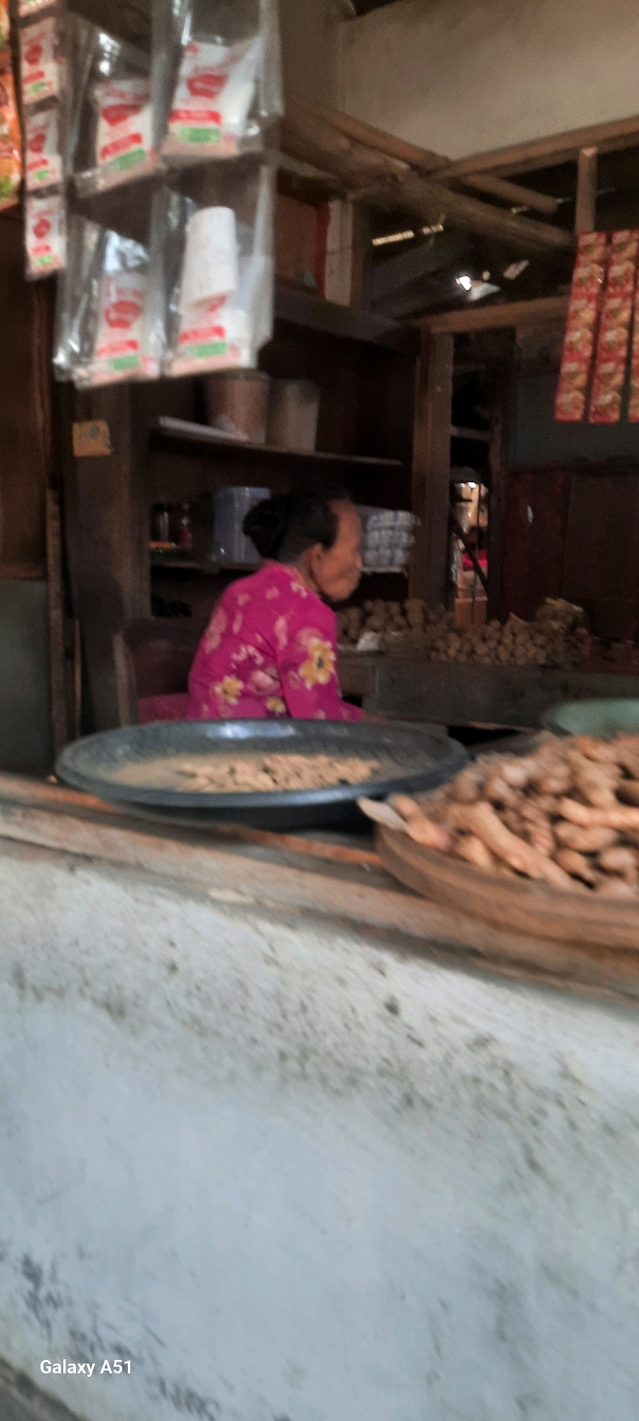 In the city of Selopuro, Indonesia, a woman with her hair pulled back in a bun is seen from the side. She is wearing a pink blouse adorned with a floral pattern. The woman is seated behind a stall. In front of her, a shallow, dark-colored circular tray holds a pile of brown, granular material. To the right of this tray is a woven basket filled with numerous long, brown, root-like objects. The stall itself appears to be an open-air market setting, with shelves and hanging bags of products visible in the background. The overall scene suggests a vendor at a local market.