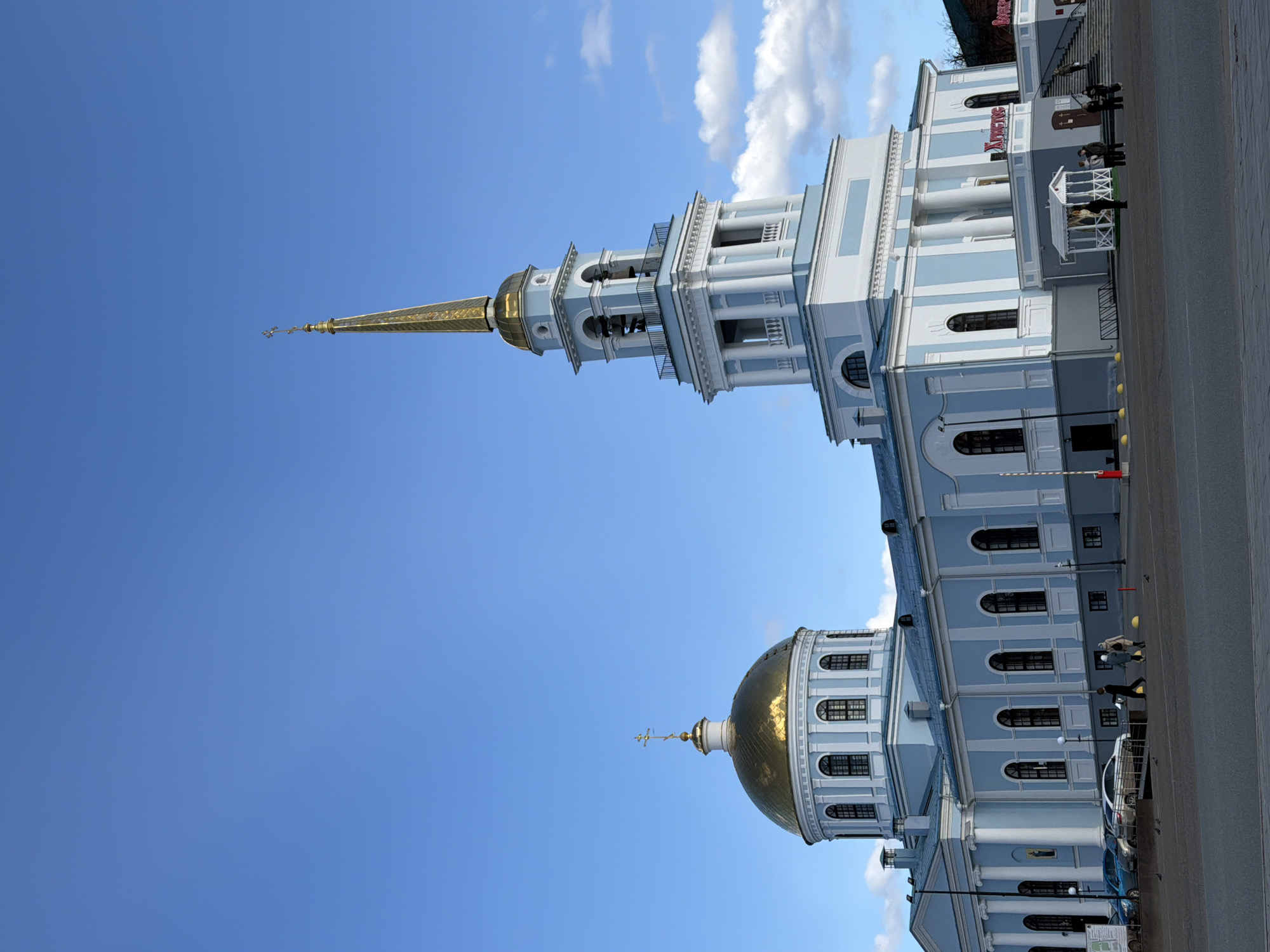 The image shows the Peter and Paul Cathedral in Votkinsk, Russia. The cathedral features a golden dome and a tall, gilded spire extending towards a bright blue sky with scattered clouds. The building is painted in light blue and white. A few people are visible in front of the building. A street runs along the right side of the frame, with parked cars and streetlights. The word "Khram" (Храм) is visible in red lettering on the facade of the building.