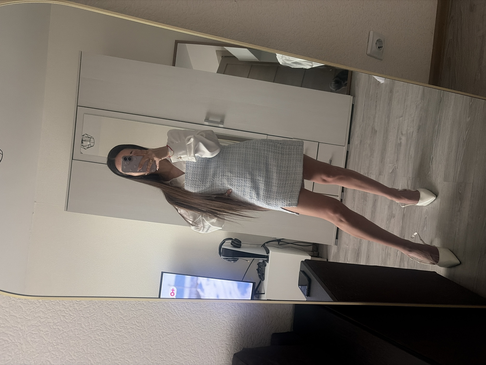 A full-length mirror selfie, taken in what appears to be an apartment in Saint Petersburg, Russia, captures a young woman posing in a modern interior. The woman, with long, straight dark brown hair, holds a smartphone with a patterned case to take the photo, partially obscuring her face. She wears a white long-sleeved blouse with cuffed sleeves and a light blue/grey textured (tweed-like) mini-dress or skirt. On her feet are white pointed-toe flat shoes with clear straps. Her left hand shows a ring, and a red string bracelet is visible on her right wrist.
The setting reflects a contemporary living space. In the background, behind the woman, are white minimalist wardrobes or cabinets with silver handles. The flooring is light-colored laminate or wood-look tiles. To the left, a television screen is visible, displaying a logo featuring a male and female symbol ("♂+♀"). Below the TV, a white stand holds a PlayStation 5 console, black over-ear headphones, and a game controller, along with various cables. Another smartphone rests on a dark brown/black low cabinet or table in the foreground. A European-style wall outlet is visible on the beige wall above the flooring on the right side of the mirror. A faint reflection of a pet, possibly a cat, can be seen in the upper right quadrant of the mirror, near the outlet. The overall impression is an organized, well-lit indoor environment.