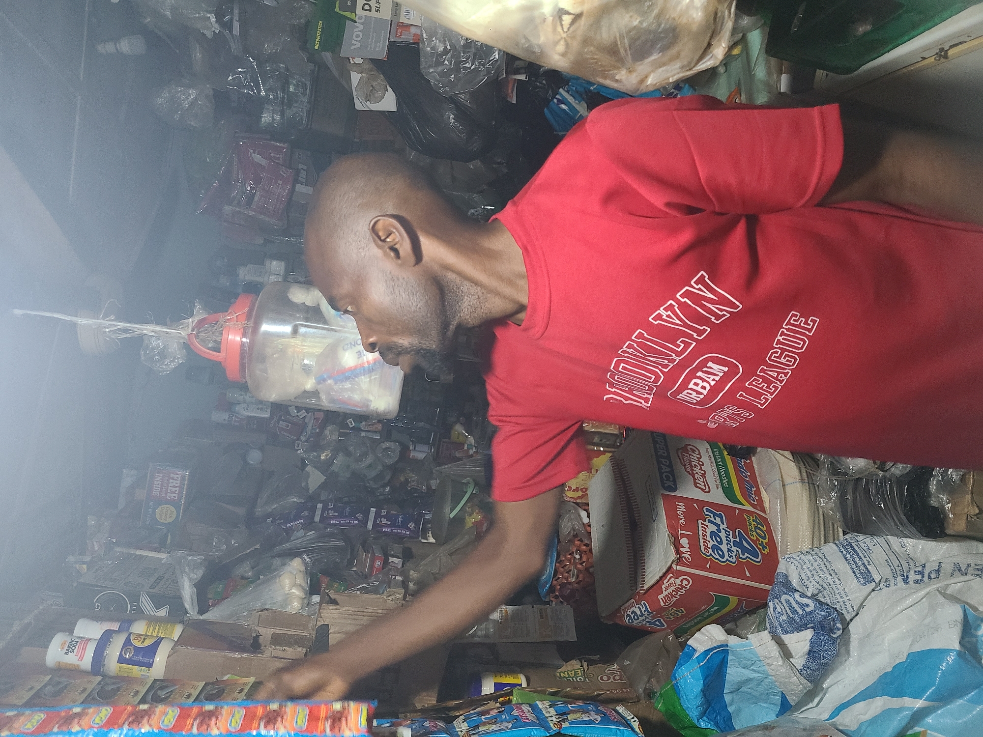 This media file features a man in a red t-shirt with white lettering that appears to say "BROOKLYN URBAM DNS LEAGUE," standing in a cluttered store or warehouse. The man is seen from the side, looking down and to his left, with a bald head and a beard. He is reaching towards something, suggesting he is in the process of picking up or arranging items.
The setting is packed with various objects, including bags, boxes, bottles, and what seem to be food products like instant noodles and possibly cleaning supplies. Some of the visible text on these items includes "FREE INSIDE," "Chicken Flavor," "VOVO DO," "MOSQUITO STICK," and nutritional information. The environment appears to be dimly lit and somewhat dusty or hazy, possibly from poor lighting or airborne particles. The overall impression is of a busy, perhaps disorganized, retail or storage space. The location context of Abuja, Nigeria, suggests an urban commercial setting. No specific time of day or weather conditions are discernible. The man's expression is neutral, focused on his task.