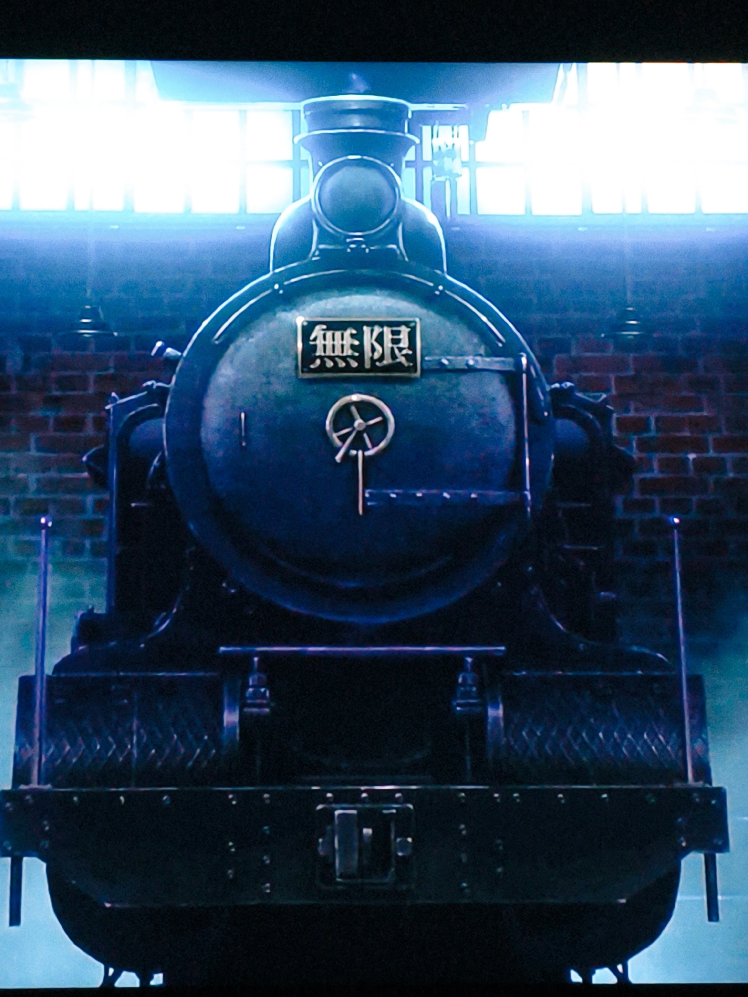 The image shows the front view of a dark-colored steam locomotive. The locomotive is predominantly black and appears to be made of metal. It has a large, circular boiler at the front, which is painted a dark blue-grey. On the boiler, there is a rectangular plate with Japanese characters that read "無限" (Mugen), meaning "infinity" or "limitless". Centered on the boiler is a circular emblem with spokes, possibly a wheel or a decorative element. Below the boiler, the locomotive has a robust frame with various mechanical components visible. There are two prominent vertical metal rods on either side of the front of the locomotive. The background consists of a brick wall and large windows or skylights that emit a bright, blue-white light, creating a stark contrast with the dark train. The overall lighting suggests an indoor or covered industrial setting. There are no people visible in the image. The image is taken from a low angle, emphasizing the imposing size of the train.
There is no indication in the image that it is located in Vladivostok, Russia. The presence of Japanese characters on the train suggests a connection to Japan or a Japanese aesthetic.