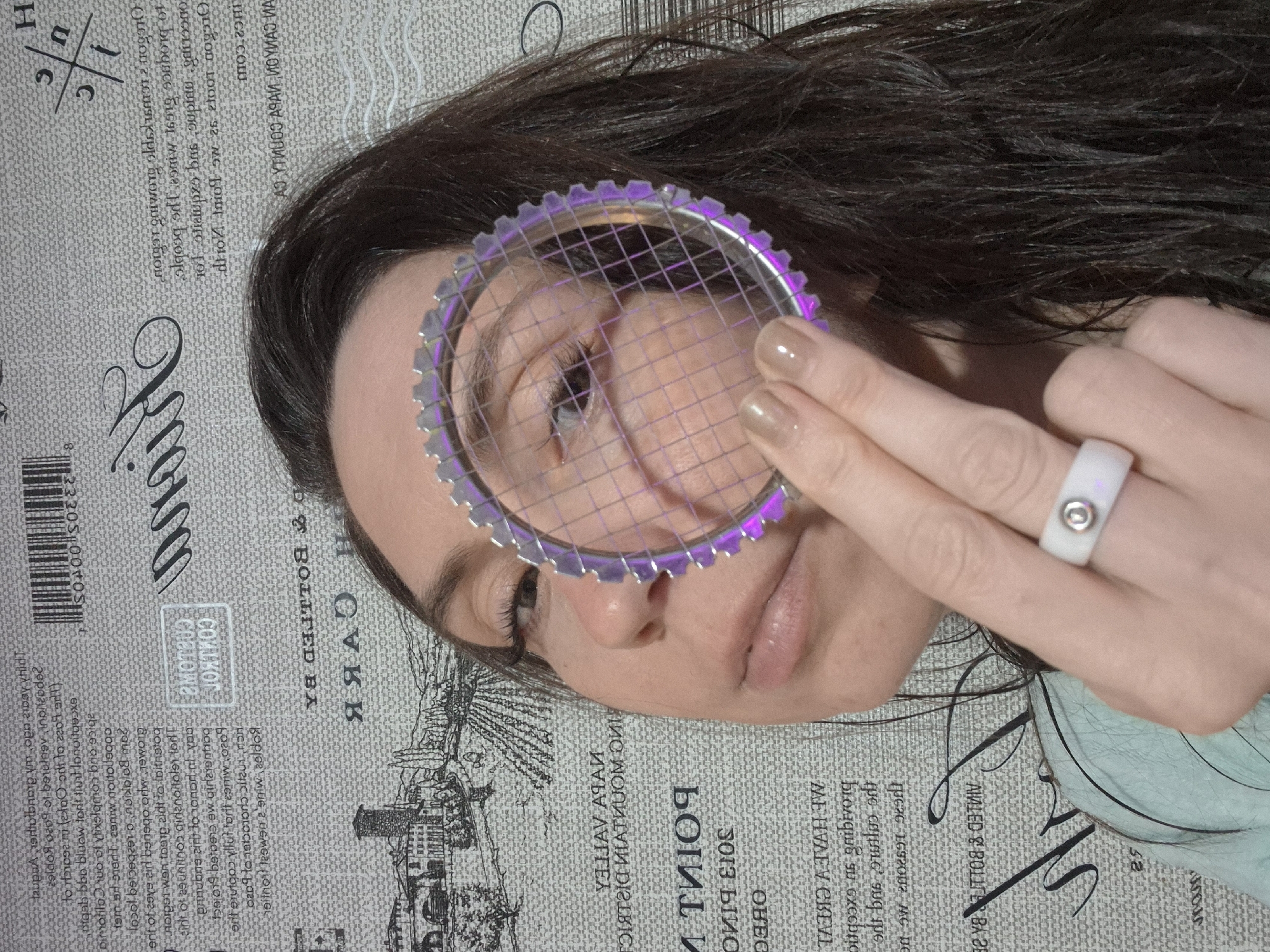A close-up, rotated photograph captures a person holding a round, purple-tinted, gear-like object in front of their eye. The individual has long, dark brown hair and light skin. The ring on their finger is white with a silver accent. The background is a gray, textured surface covered in what appears to be newspaper print, featuring text and illustrations of buildings. The overall lighting is diffused, casting soft shadows and highlighting the texture of the surface.