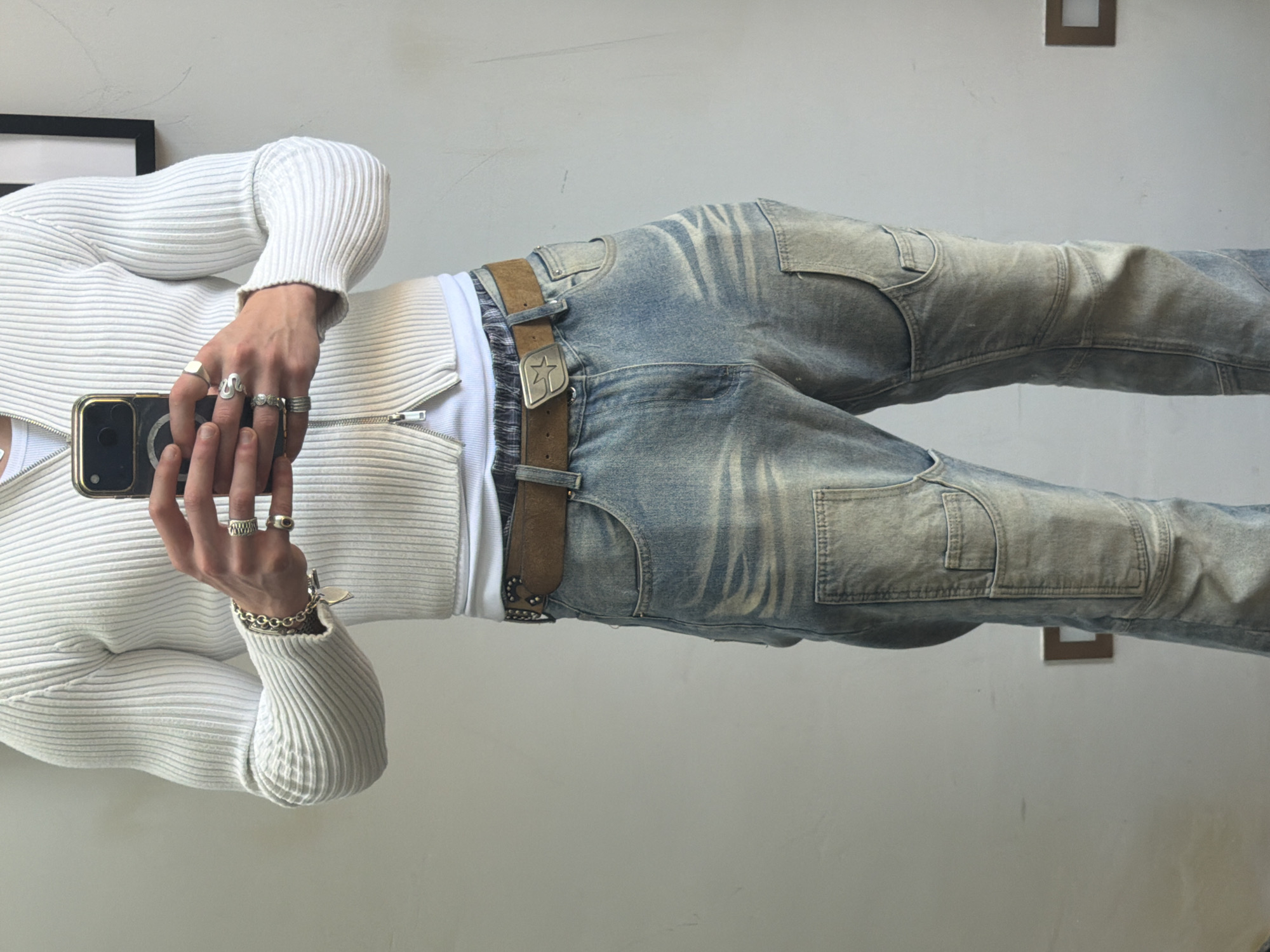 A person wearing a white ribbed cardigan, white t-shirt, and faded blue jeans with cargo pockets poses for a mirror selfie. They are holding a gold-colored mobile phone in their hands, which are adorned with multiple rings and a gold bracelet. The jeans are secured with a brown leather belt featuring a star-shaped buckle. The background includes a framed picture and a decorative wall hanging. The location and specific details related to London are not discernible from the image.
