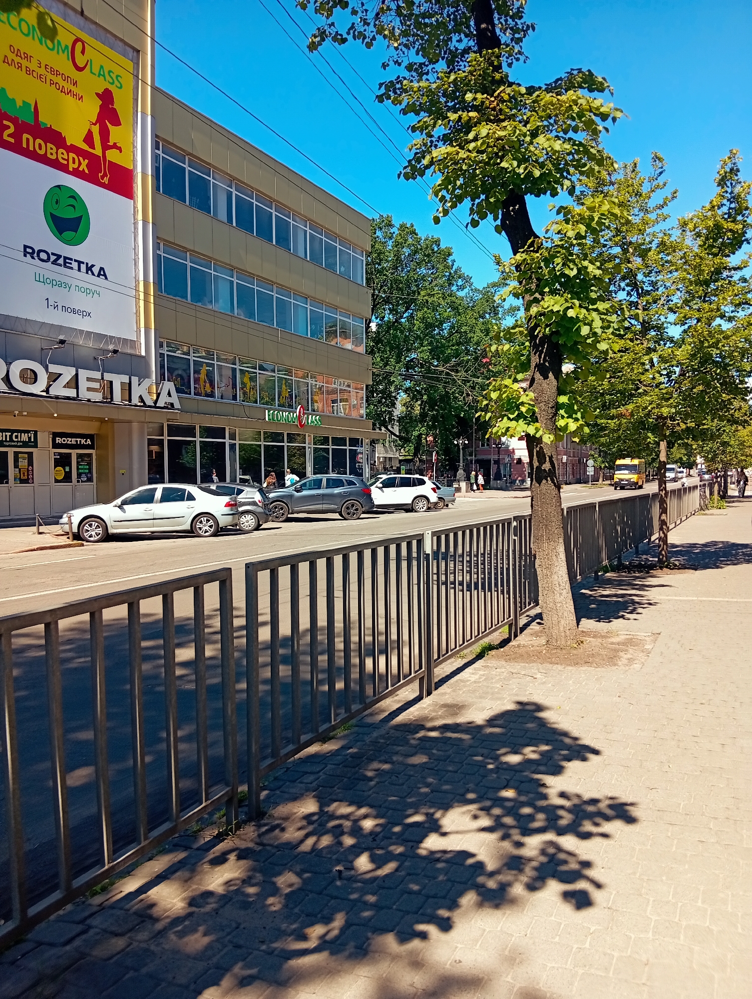 This image captures a vibrant, sunlit urban street scene in Poltava, Ukraine, likely during mid-morning or early afternoon due to the strong shadows cast by trees and a clear blue sky.
In the foreground, a dark metal fence separates the street from a paved sidewalk on the right. Tall green trees with dense foliage line the sidewalk, their branches creating intricate shadows on the light-colored pavers.
The left side of the image is dominated by a multi-story commercial building featuring large, reflective windows. Prominent signage indicates the presence of a "ROZETKA" pickup point, a well-known Ukrainian online retailer, with its logo displayed multiple times. A large banner on the building advertises "ECONOM CLASS," stating "ОДЯГ З ЄВРОПИ ДЛЯ ВСІЄЇ РОДИНИ" (Clothes from Europe for the whole family) and indicating that it is on the "2 поверх" (2nd floor). The ROZETKA sign also mentions "Щоразу поруч" (Always nearby) and "1-й поверх" (1st floor). Other visible text fragments include "ВІТ СІМ'Ї" and "торговий дім" (shopping mall/department store).
Several cars are parked along the street in front of the building, including a white sedan, a grey hatchback, and a grey SUV. A yellow van is also visible further down the road. A few pedestrians can be seen walking on the sidewalks in the distance, engaging in daily urban activity. The overall setting is a bustling commercial area, characterized by retail establishments, traffic, and pedestrian movement under bright, clear weather conditions.