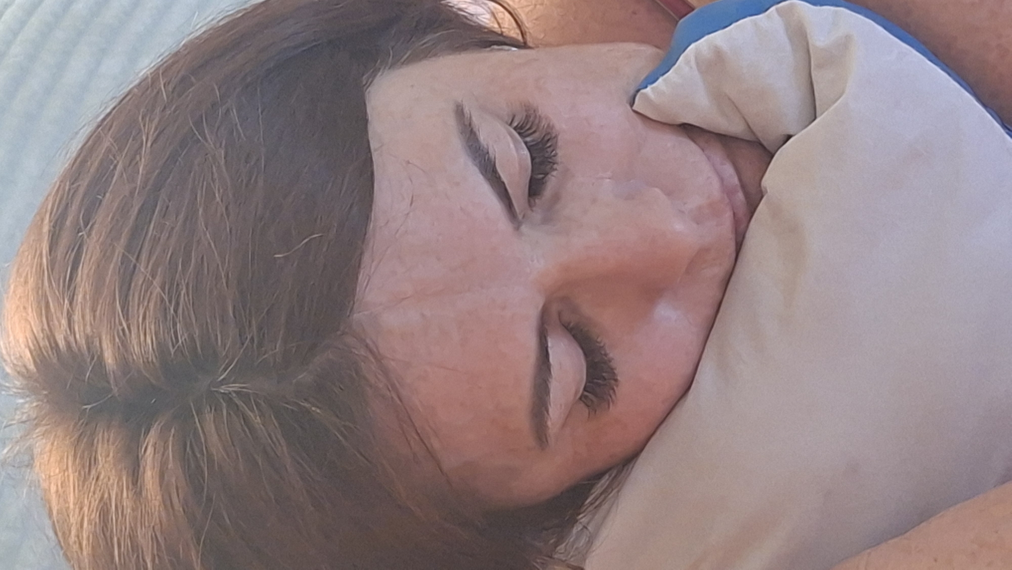 A close-up, overhead shot shows a woman with auburn hair resting her head on a light-colored pillow. Her eyes are closed, and she appears to be sleeping or resting. The woman has long, dark eyelashes, and subtle freckles are visible on her forehead. She is wearing a pink top. The background is a muted blue. The image does not provide any contextual information about the location in Voinova, Moldova.