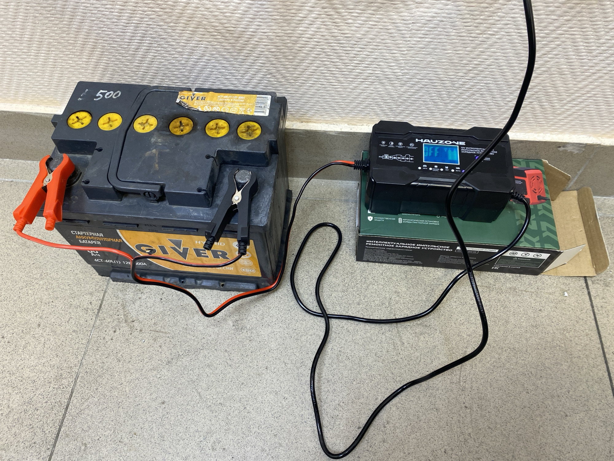The image shows a GIVER car battery, model 6CT-60L(1) 12V 500A, connected to a Hauzone battery charger. The battery charger is sitting on its original packaging box. The battery has a handwritten "500" on the top. The scene appears to be indoors on a tiled floor against a wall. The text on the battery indicates it was made in Russia, while the charger box indicates it was made in China. There is no indication of the city of Sochi in this image.